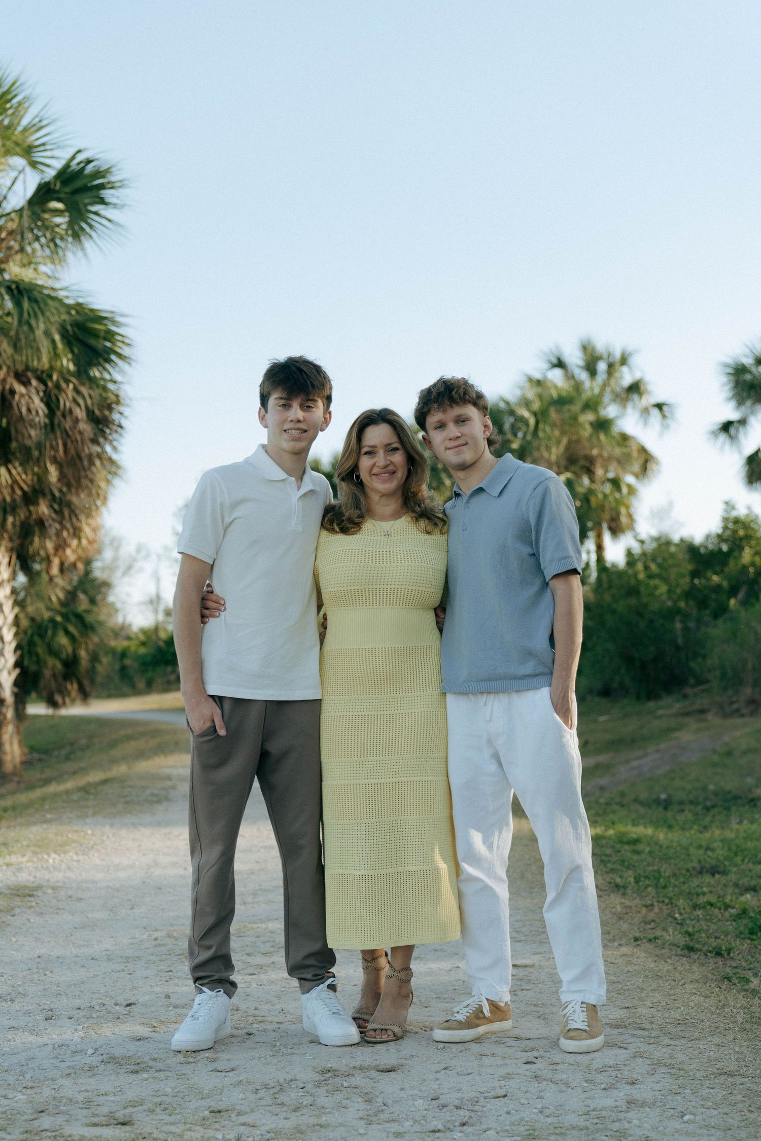 Three people stand together on a dirt path with palm trees in the background during golden hour lighting.