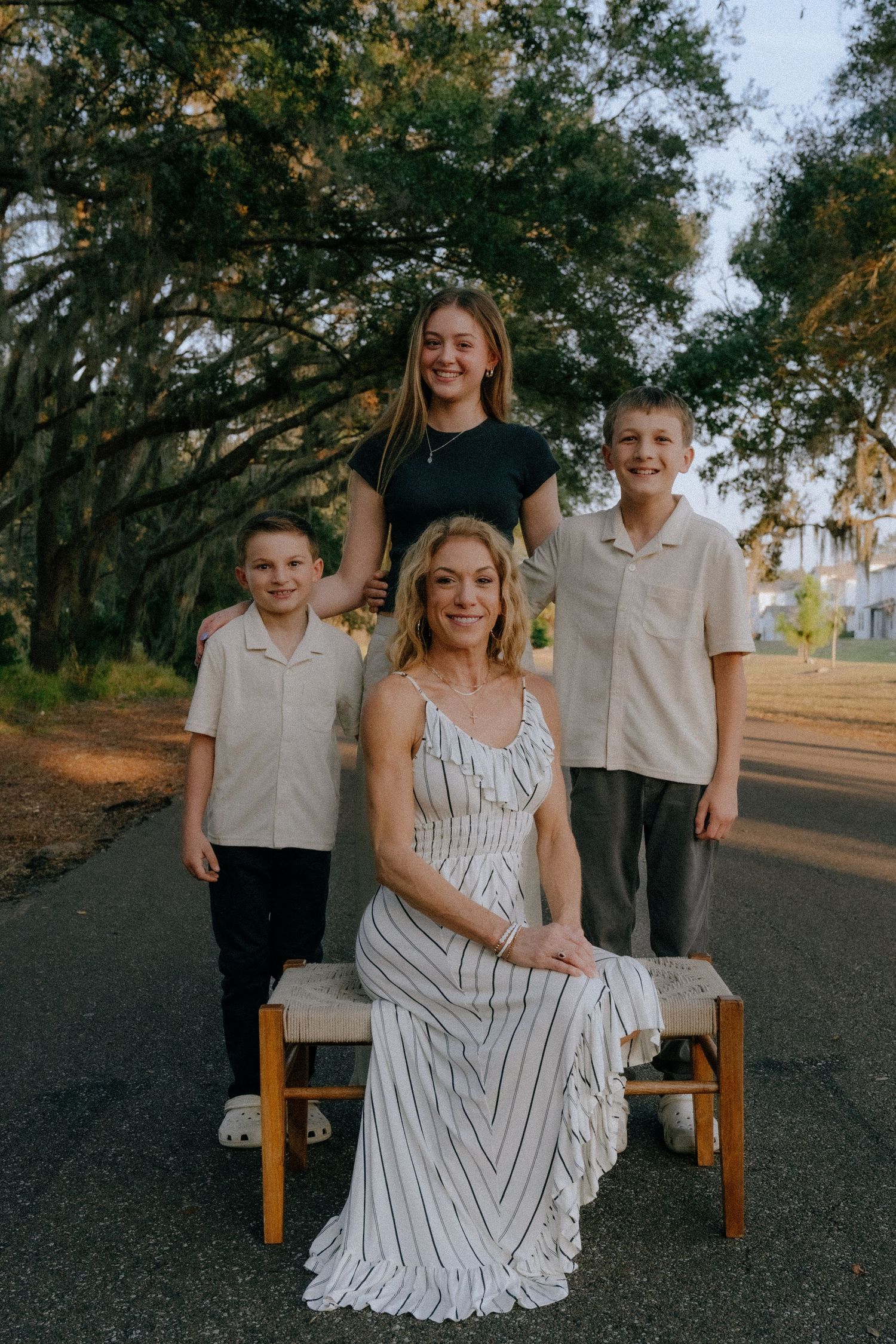 A family poses together on a wooden bench along a tree-lined path during an outdoor photo session.