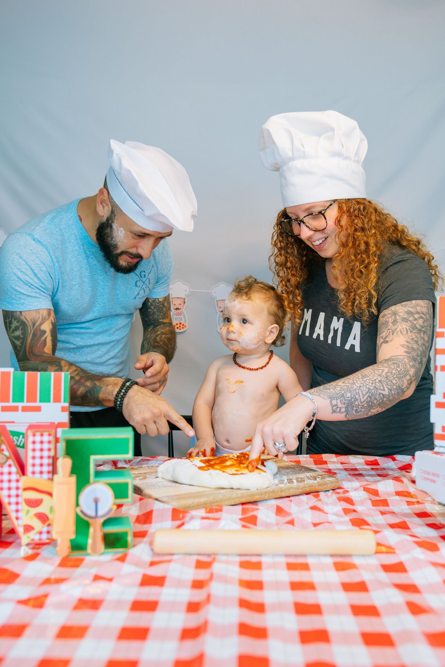 Family wearing chef hats prepares food together at table with red checkered tablecloth in home kitchen setting.