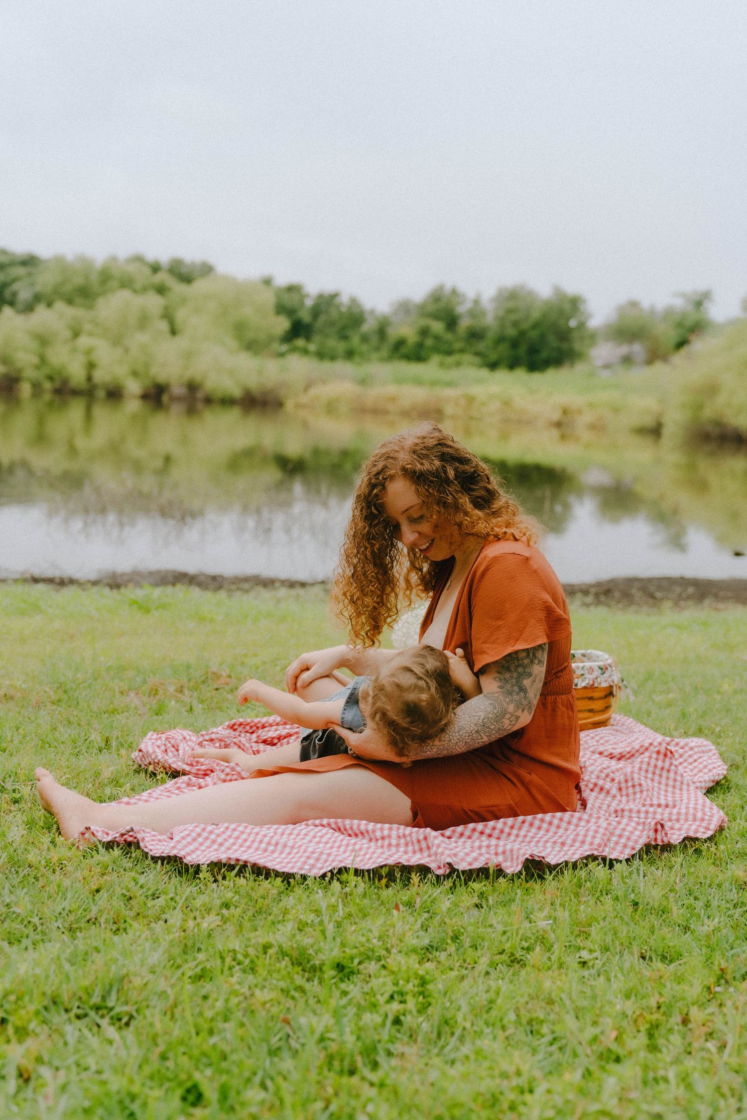 A peaceful afternoon picnic by a pond with a red gingham blanket spread on the grass.