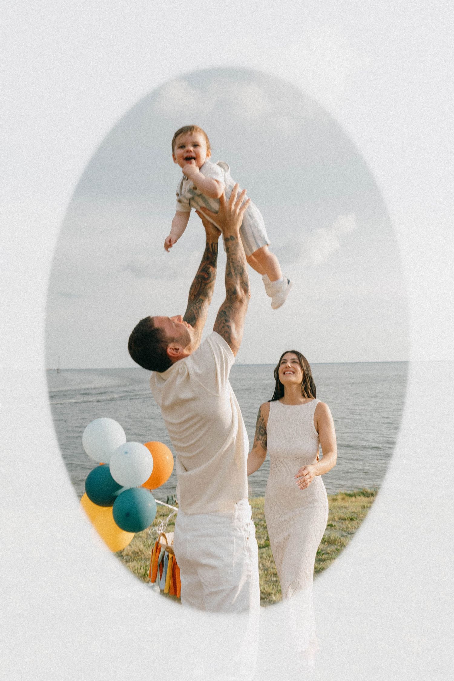 Family celebrating outdoors by the sea, father lifting baby in the air while mother smiles, with colorful balloons nearby.