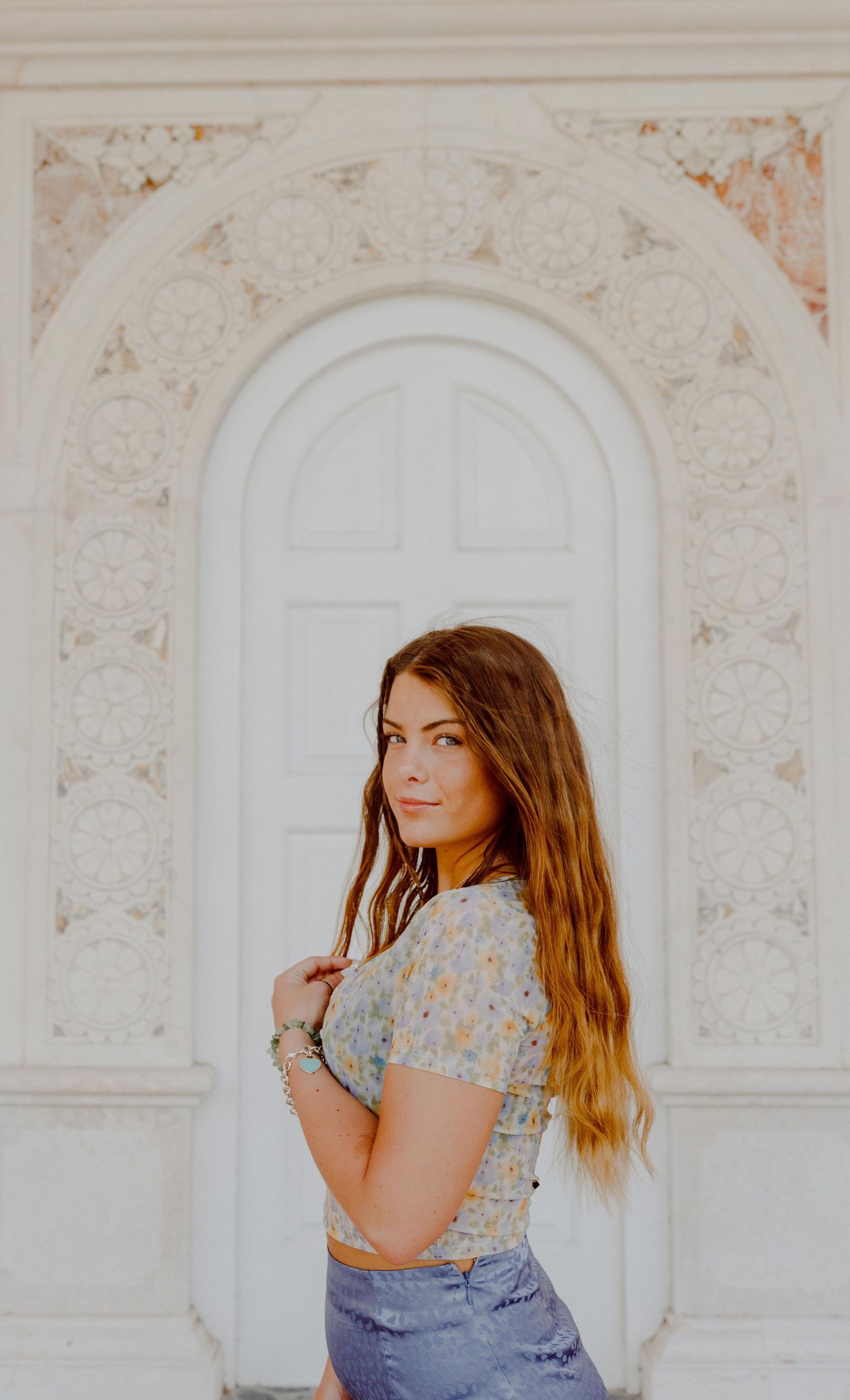 A person in floral top and jeans poses in front of an ornate white arched doorway.