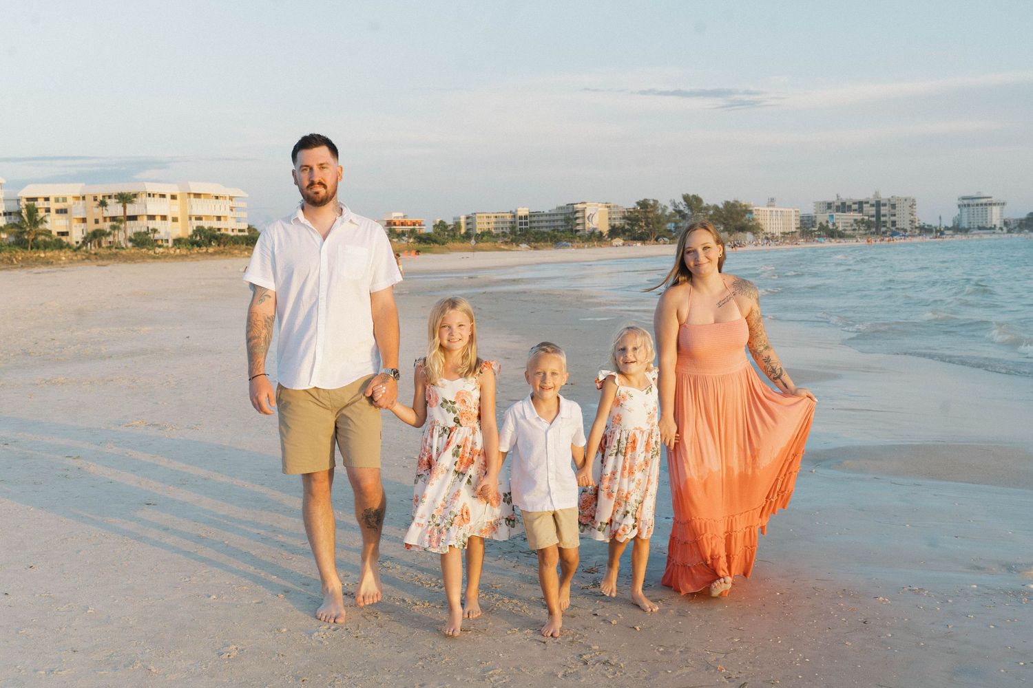 Family of five walking hand-in-hand on a sunny beach, parents and three children in coordinating outfits.
