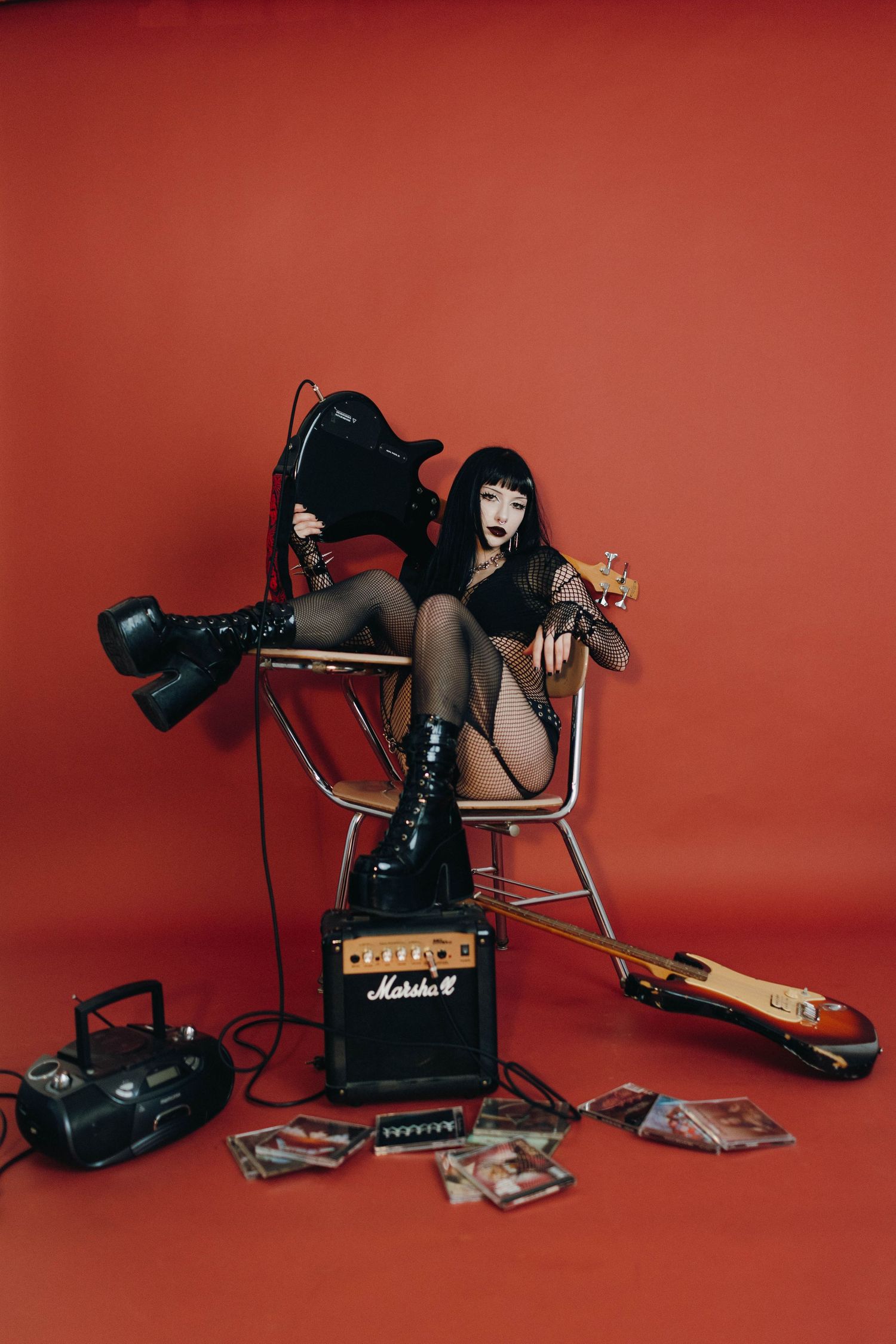 Goth woman in black outfit sits on chair with Marshall amp, electric guitars, and boombox on red background.