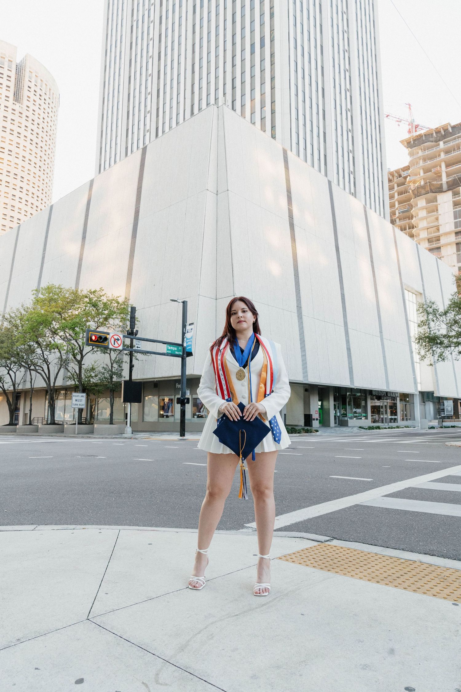 Young woman in white blazer and shorts wearing a medal stands confidently at urban crosswalk with skyscrapers behind her.