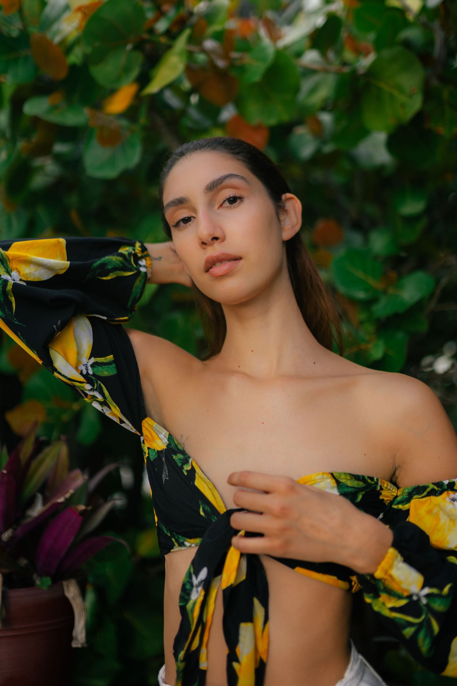 Portrait against lush green foliage wearing a black and yellow floral off-shoulder dress in warm afternoon light.