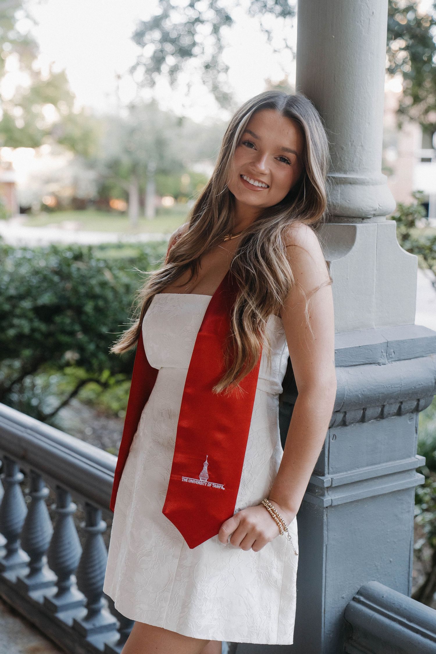 Smiling young woman in white dress wearing red graduation sash poses on decorative balcony outdoors.