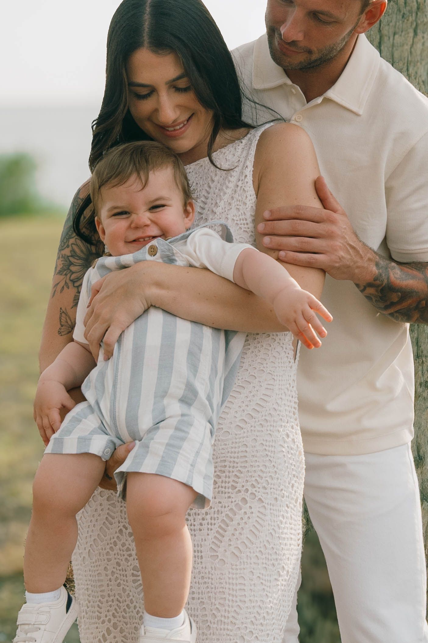 Happy young family with tattooed parents holding smiling baby outdoors near tree in white summer outfits.