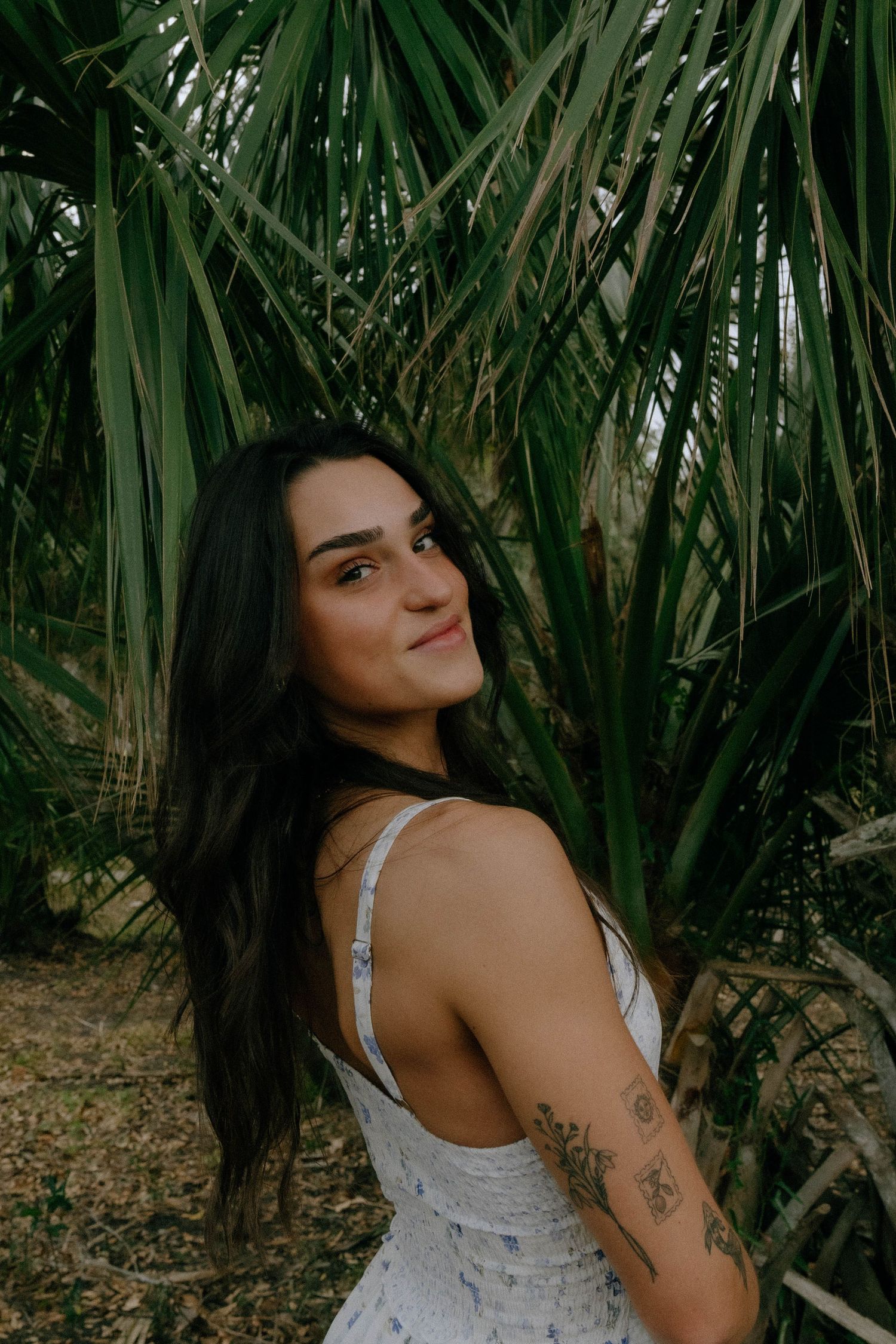 A person in a white sundress poses against tropical palm fronds in a garden setting.