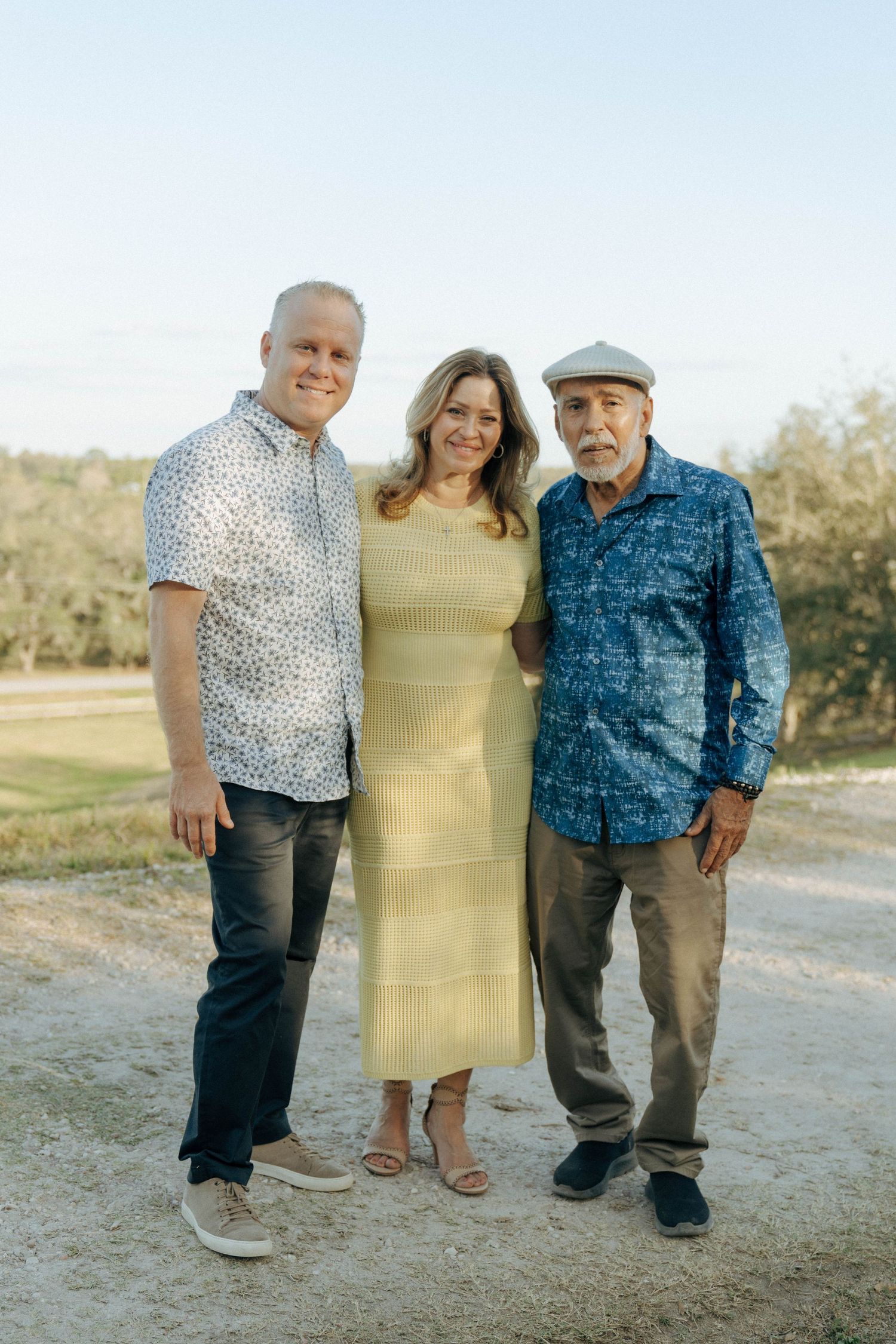 Three people pose together outdoors on a dirt path, wearing casual attire in warm evening sunlight.