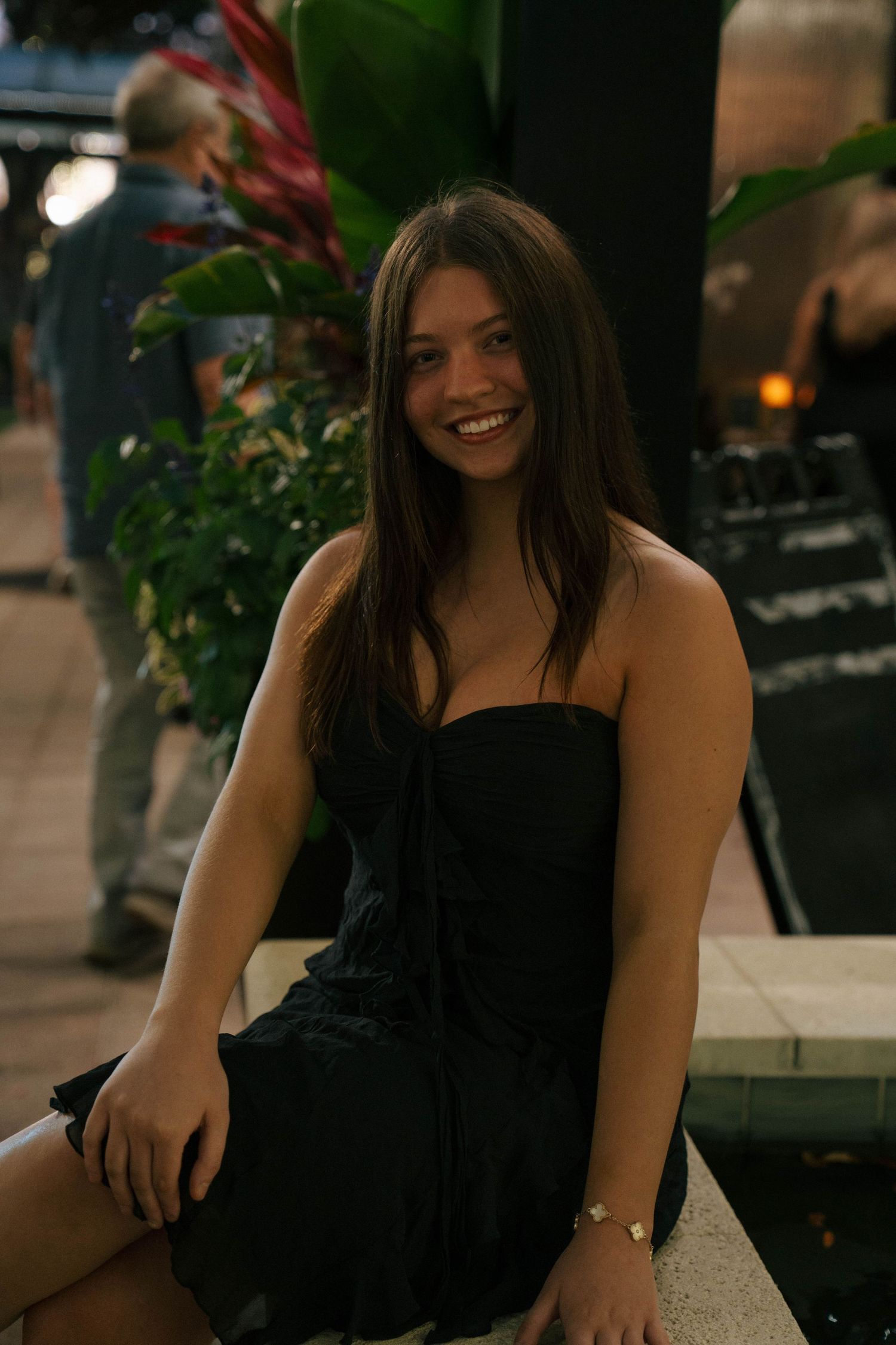 A person in a black strapless dress sits casually on a stone ledge with greenery and soft lighting in the background.