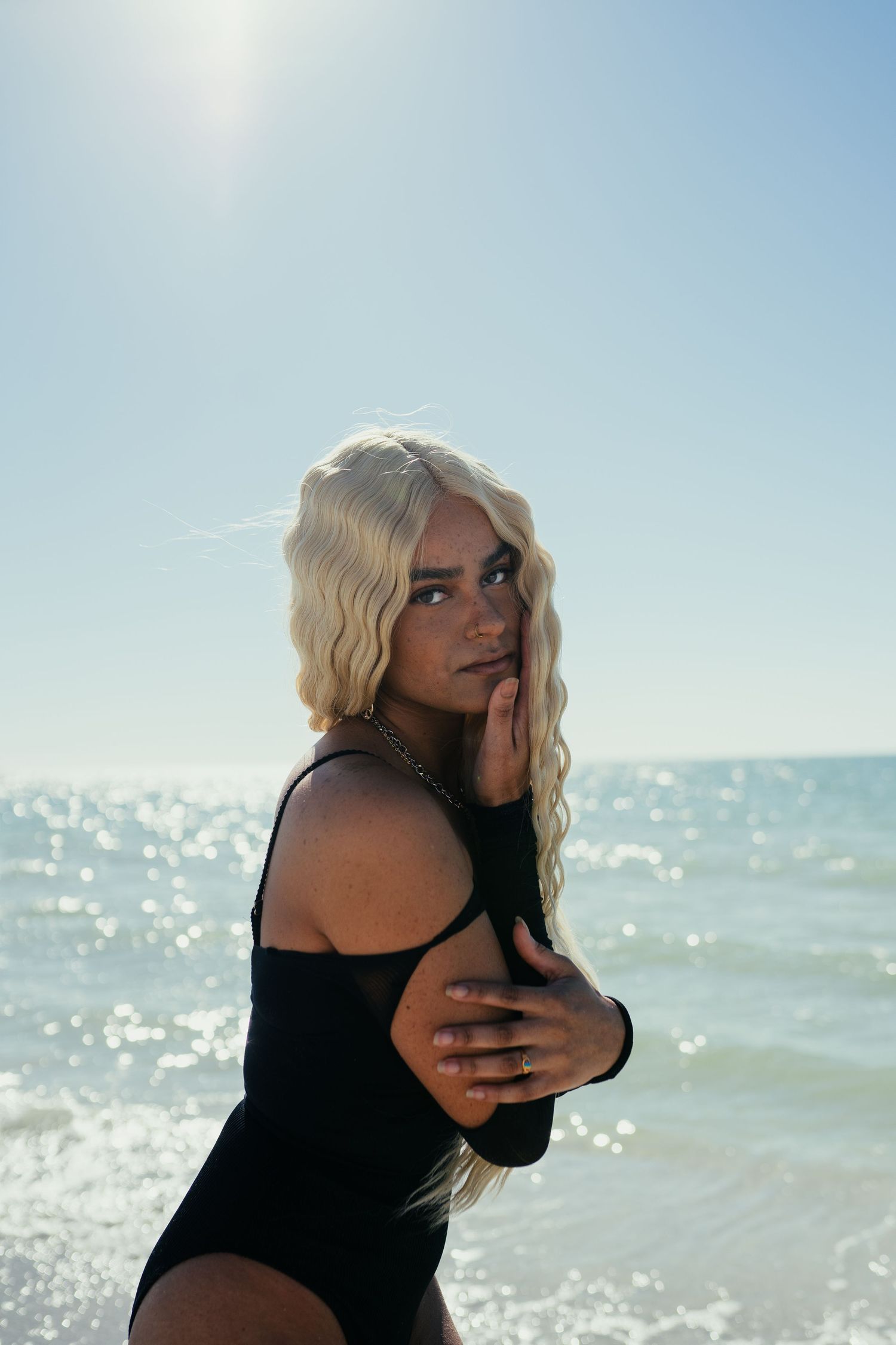 A person in a black swimsuit poses on a sunny beach with sparkling ocean waves and blue sky in the background.