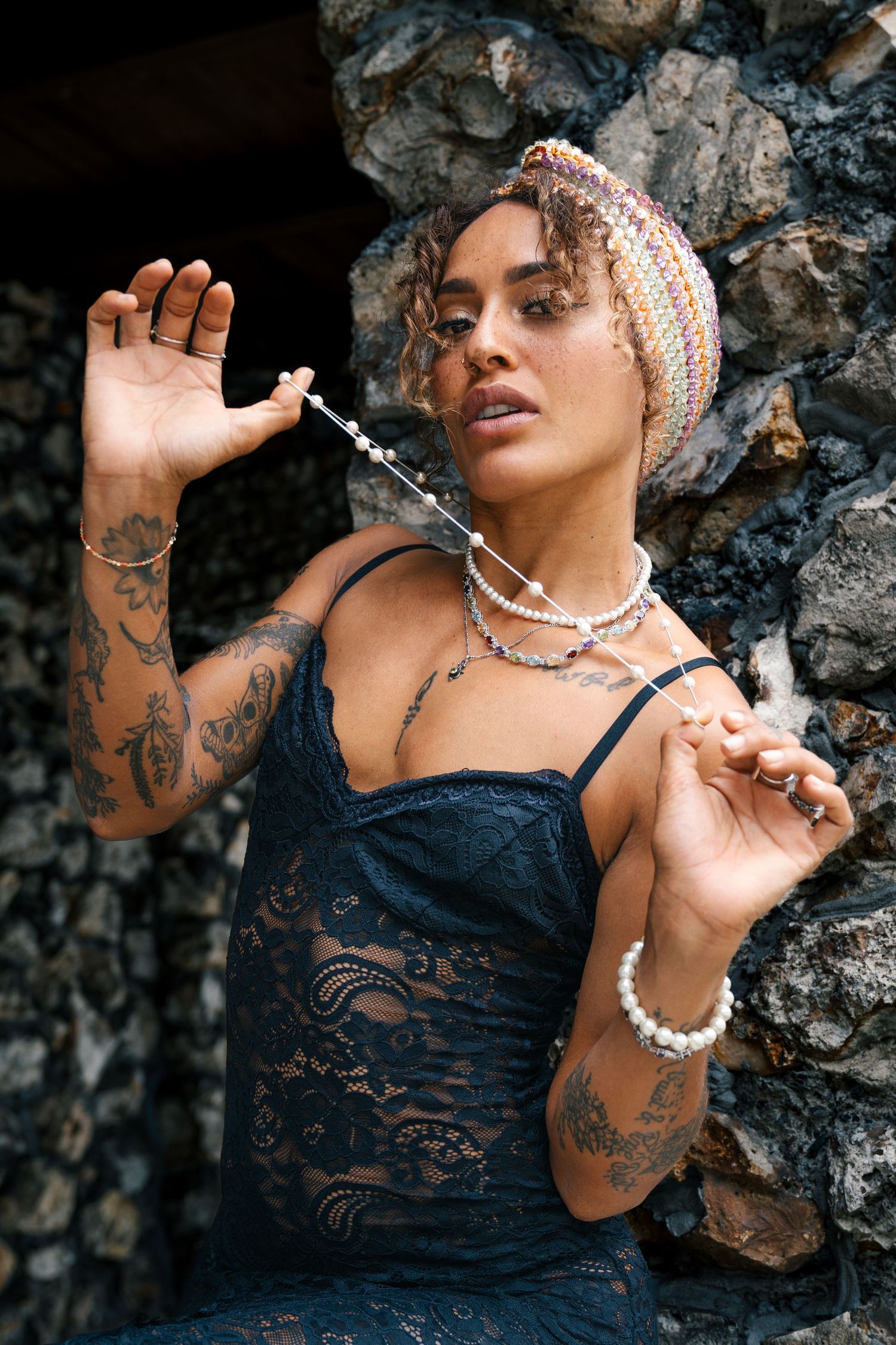 Artistic portrait against stone wall featuring lace dress, beaded pearl necklace, head wrap, and decorative tattoos.