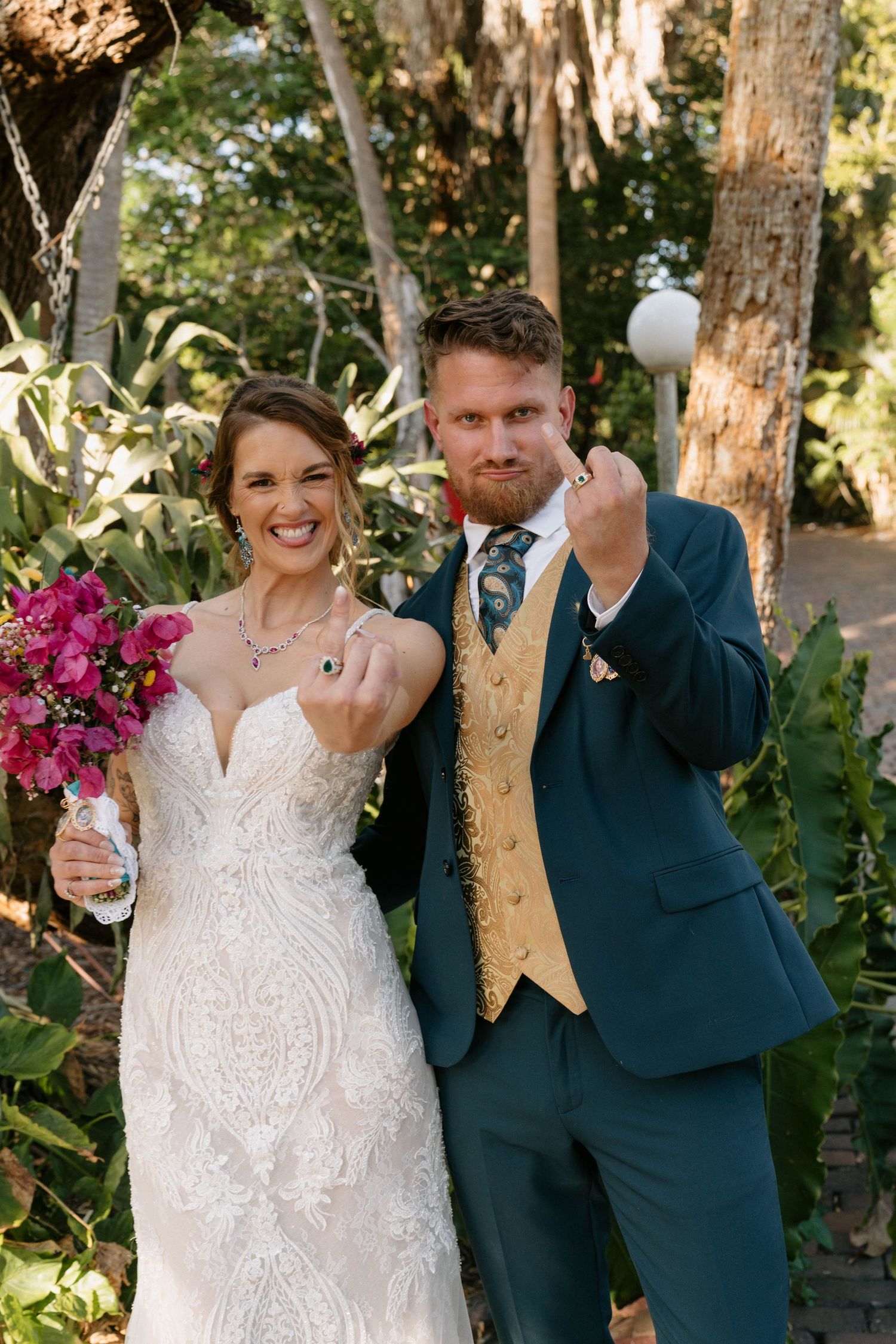 Newlywed couple showing off wedding rings outdoors, bride in lace gown with pink bouquet, groom in teal suit with gold vest.