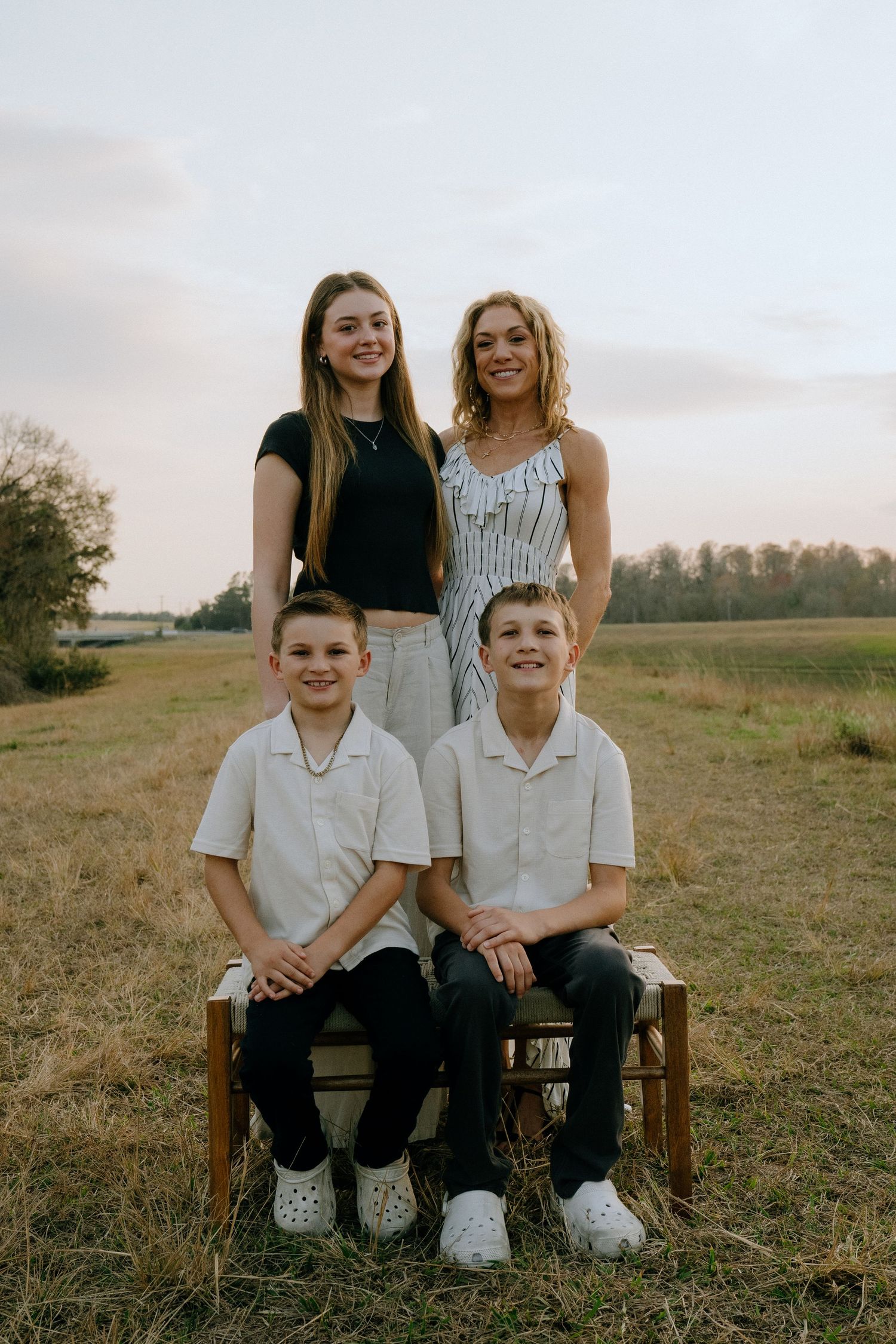 A family portrait in a rustic outdoor setting at sunset with two seated on a wooden bench and two standing behind.