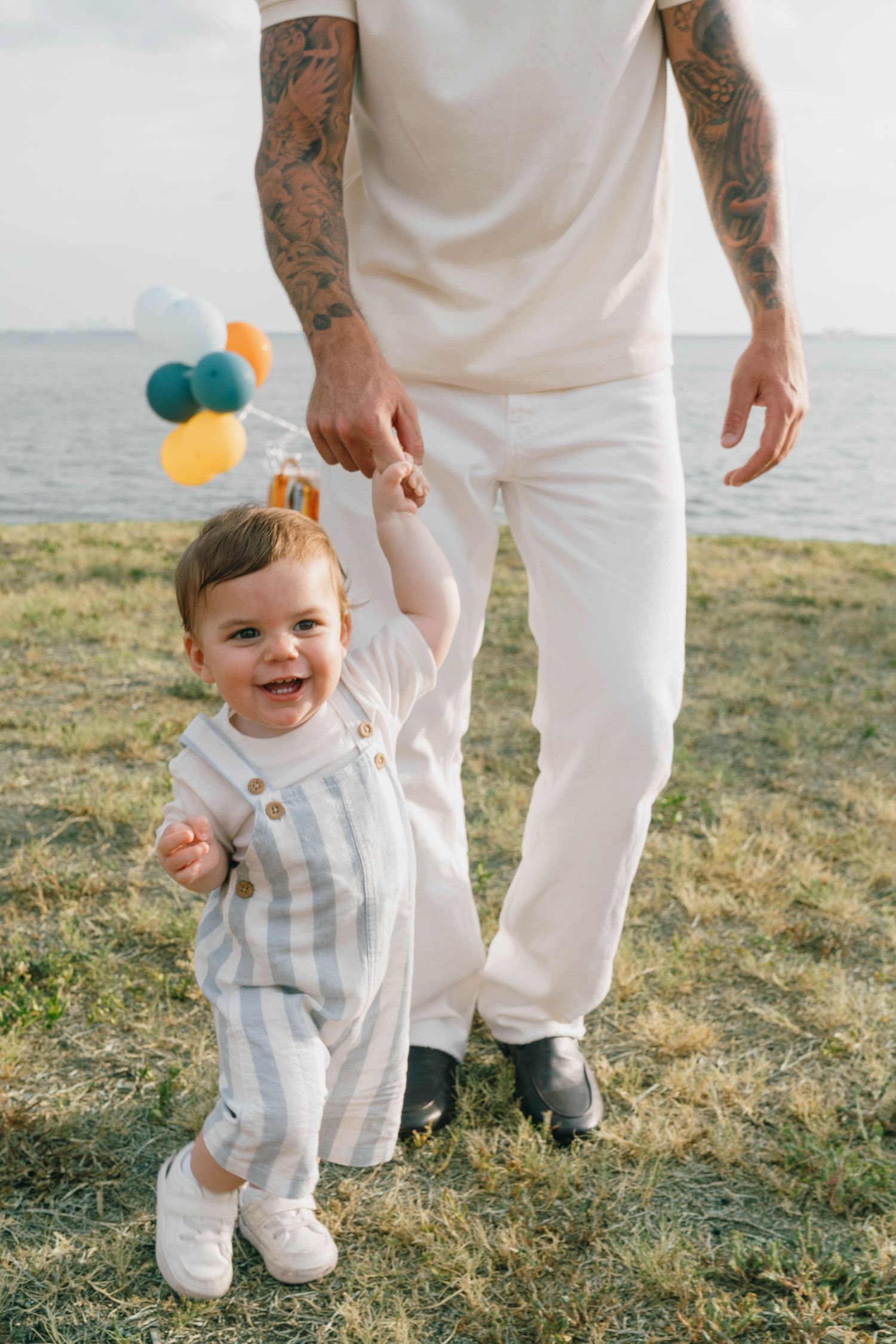 Tattooed father holds hands with smiling baby holding colorful balloons, walking on grass near the ocean.