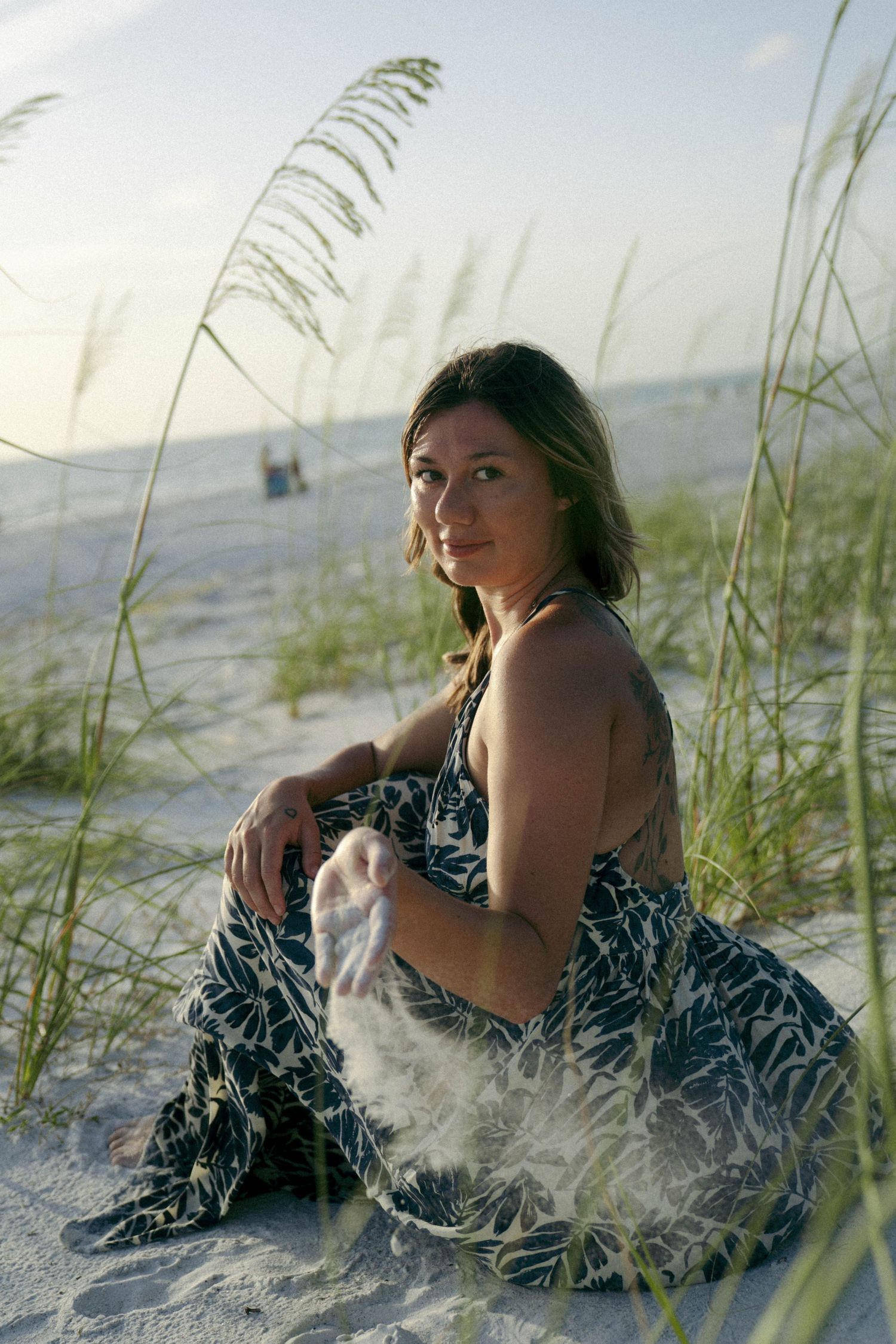 A woman in a patterned dress poses on a sandy beach with sea grass and ocean waves in the background.