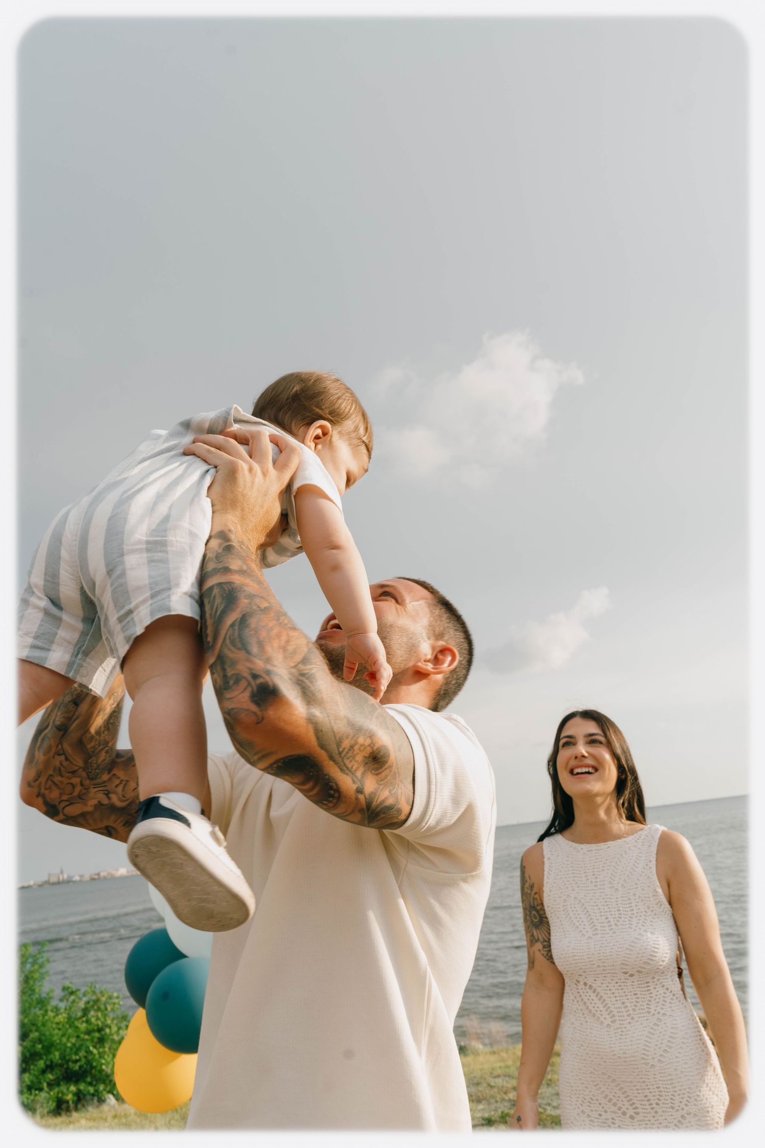 A tattooed father lifts his baby boy while a smiling woman in white dress stands nearby at a seaside outdoor celebration.