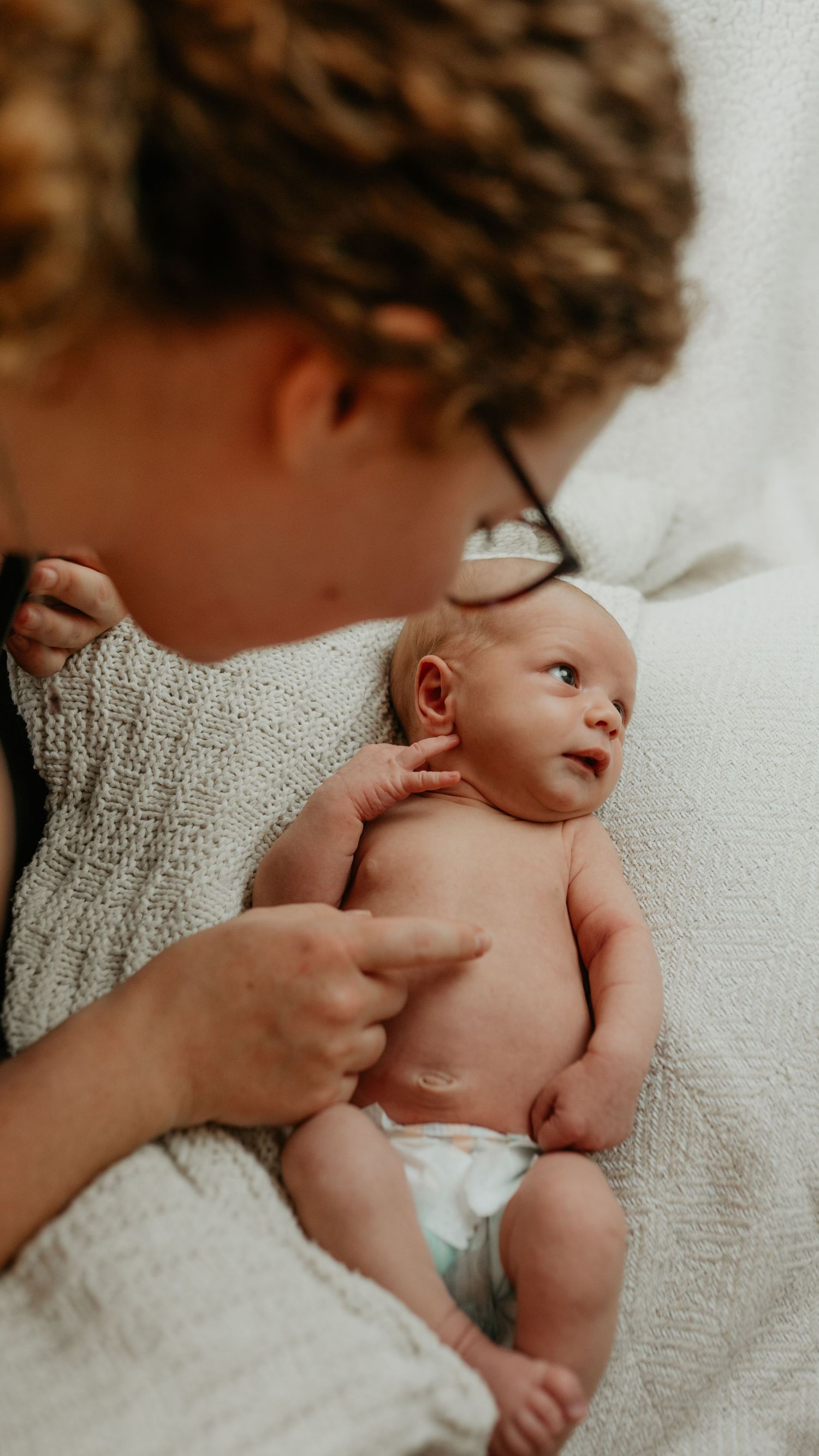 A tender moment between caregiver and newborn baby lying on soft white blankets in warm, natural lighting.
