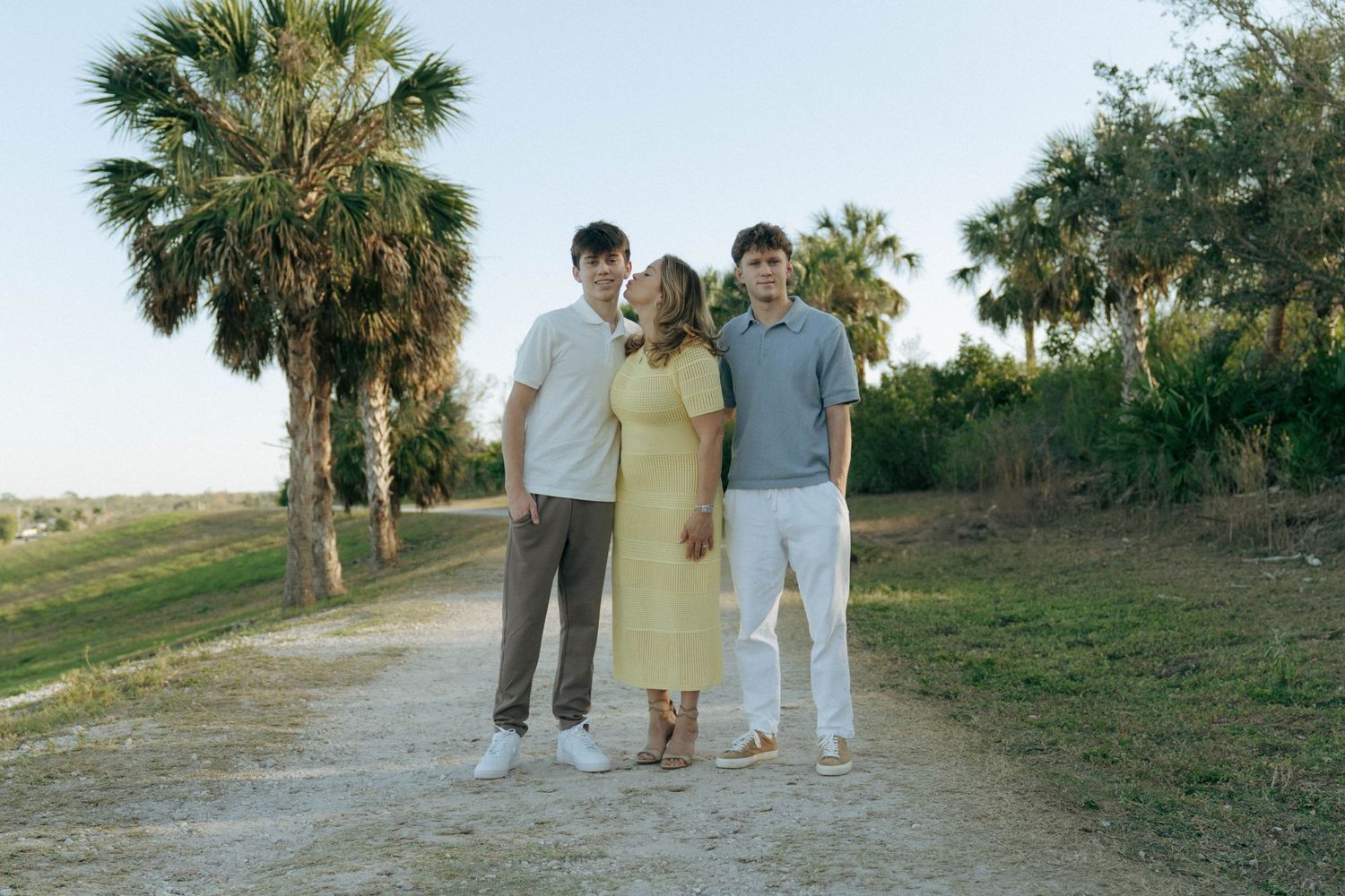Three people stand together on a dirt path with palm trees and green foliage in the background on a sunny day.