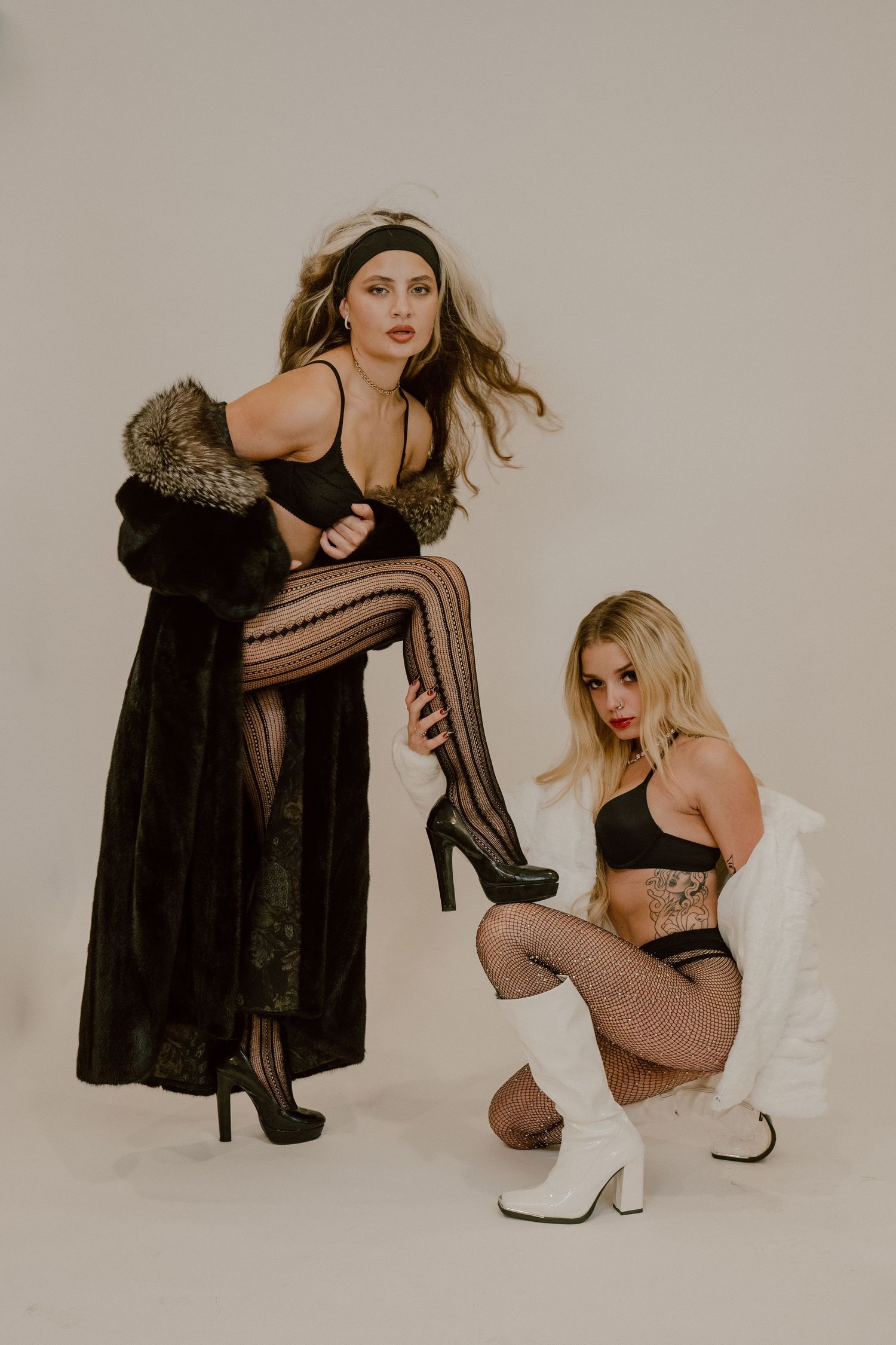 Two models pose in dark and white fur coats with fishnet stockings and platform boots against a neutral studio backdrop.