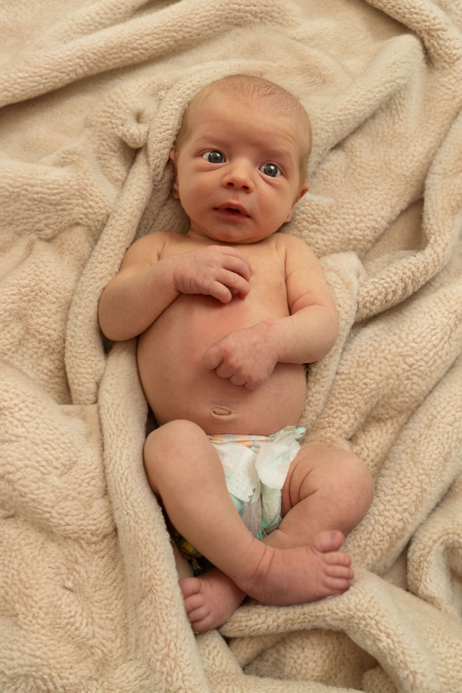 A newborn baby wearing a diaper lies on a soft beige blanket while looking up with wide eyes.