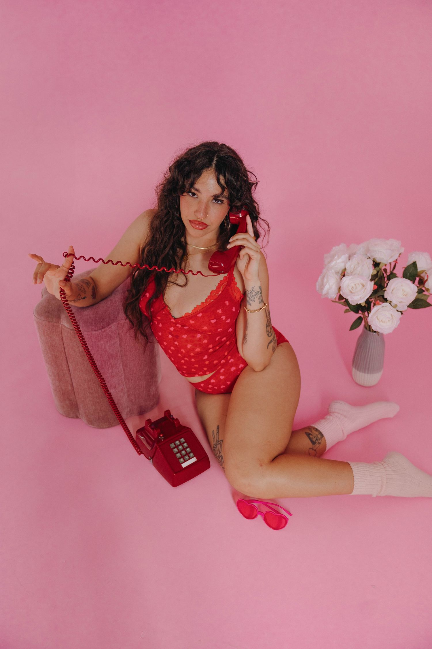Woman in red bodysuit poses with vintage red telephone against pink background with flowers.