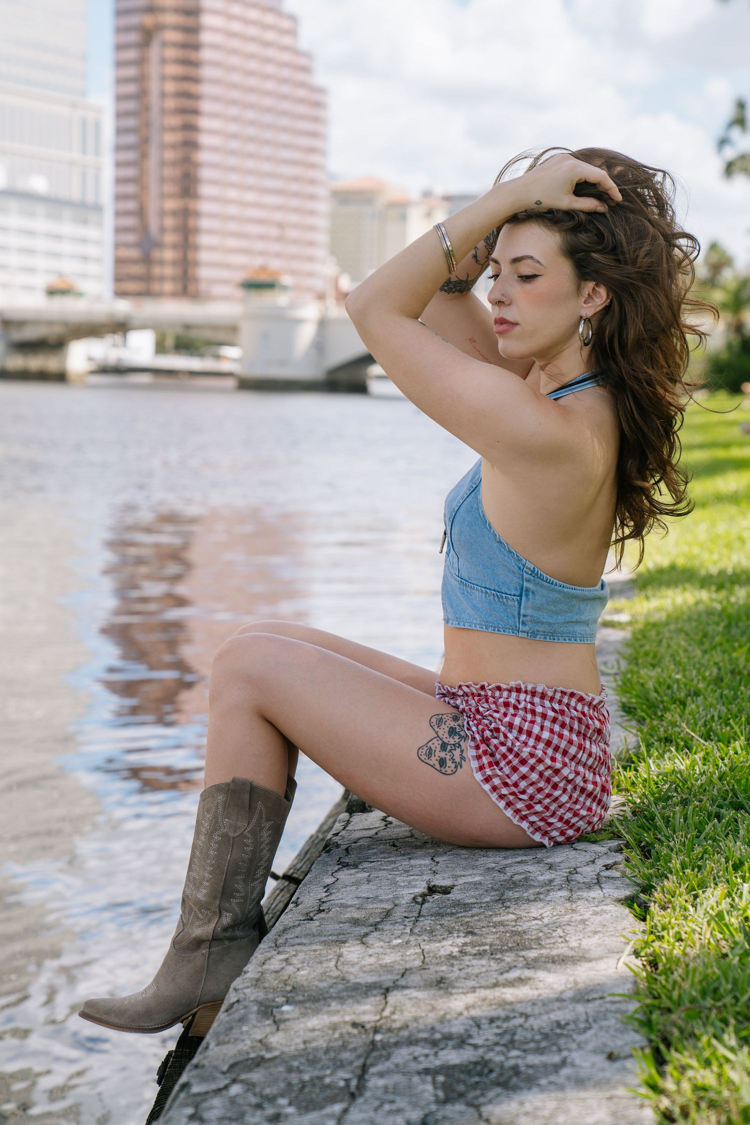A woman in a denim crop top and patterned shorts relaxes by a riverfront with city buildings in the background.
