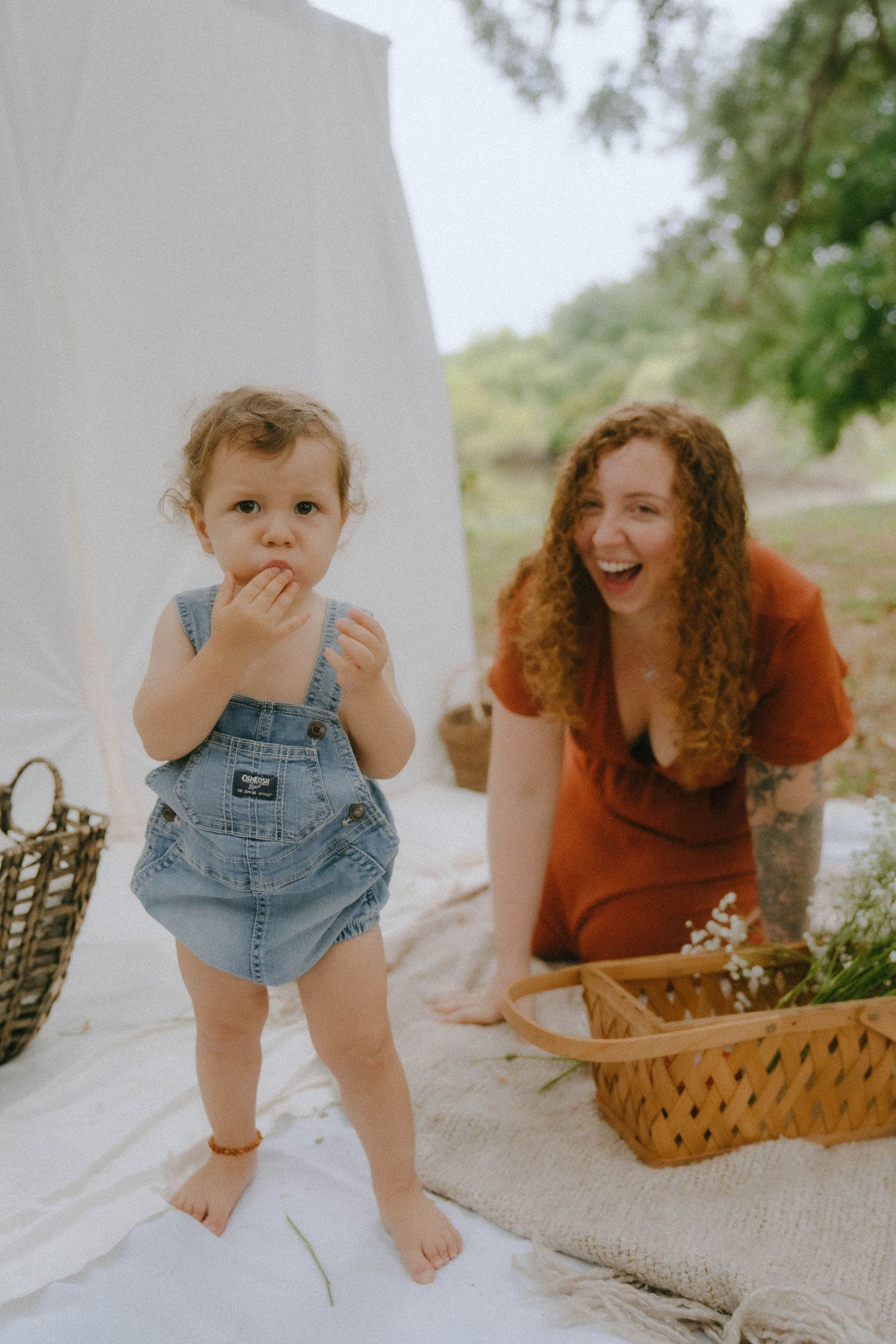 A picnic scene with a toddler in denim overalls standing on a white blanket next to a wicker basket.