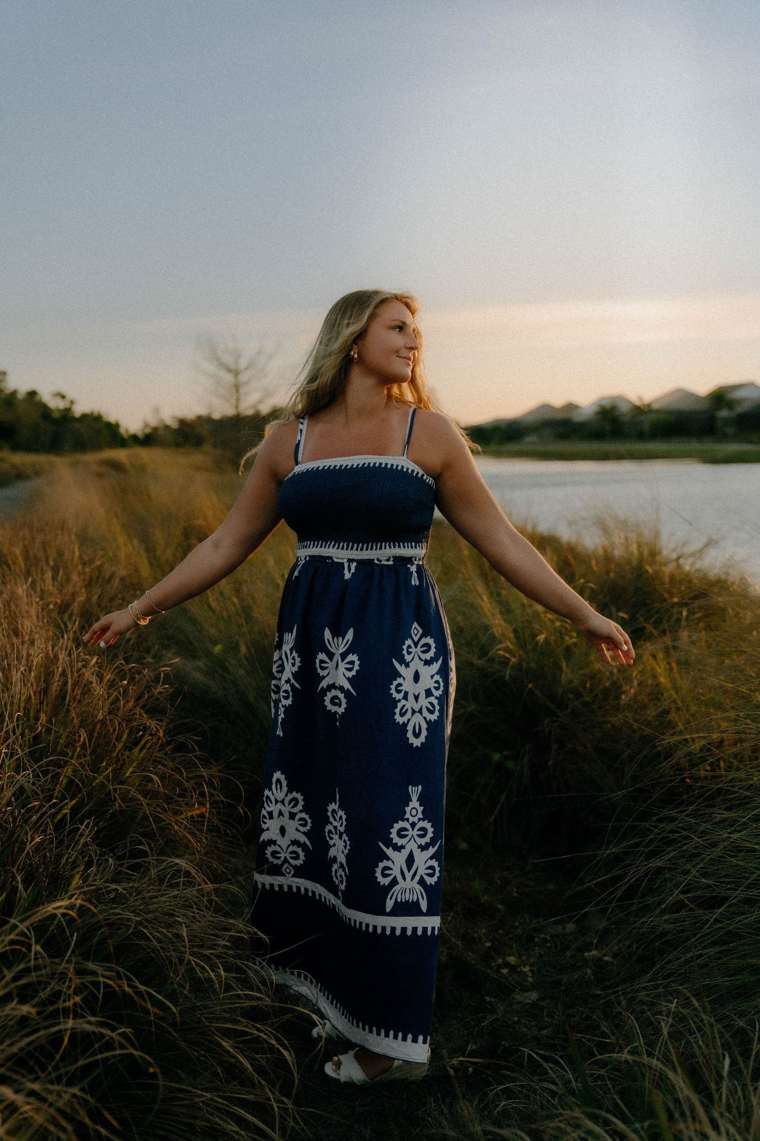 A woman in a navy and white patterned maxi dress stands in tall grass by a lake at sunset.