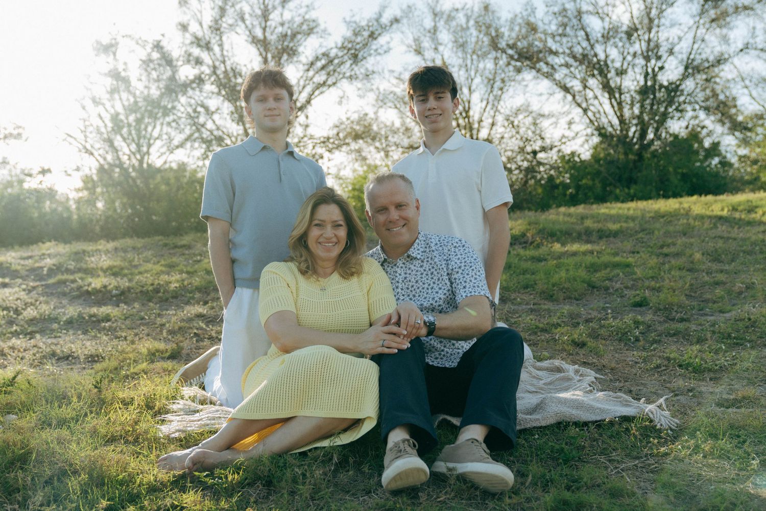 Four people sitting together on a blanket outdoors during golden hour with trees in the background.
