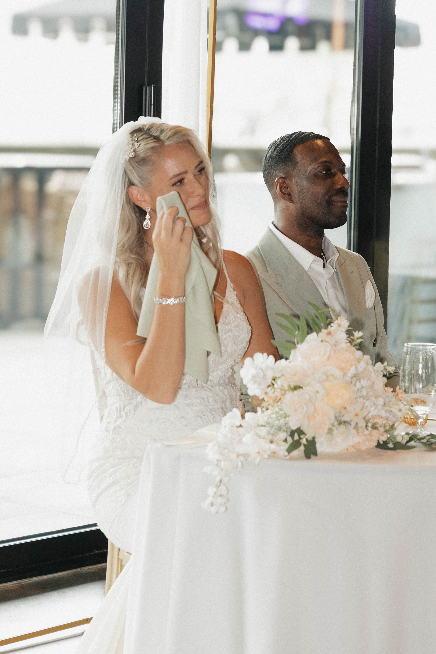 Emotional bride wiping tears at wedding reception sweetheart table with groom in beige suit beside her.
