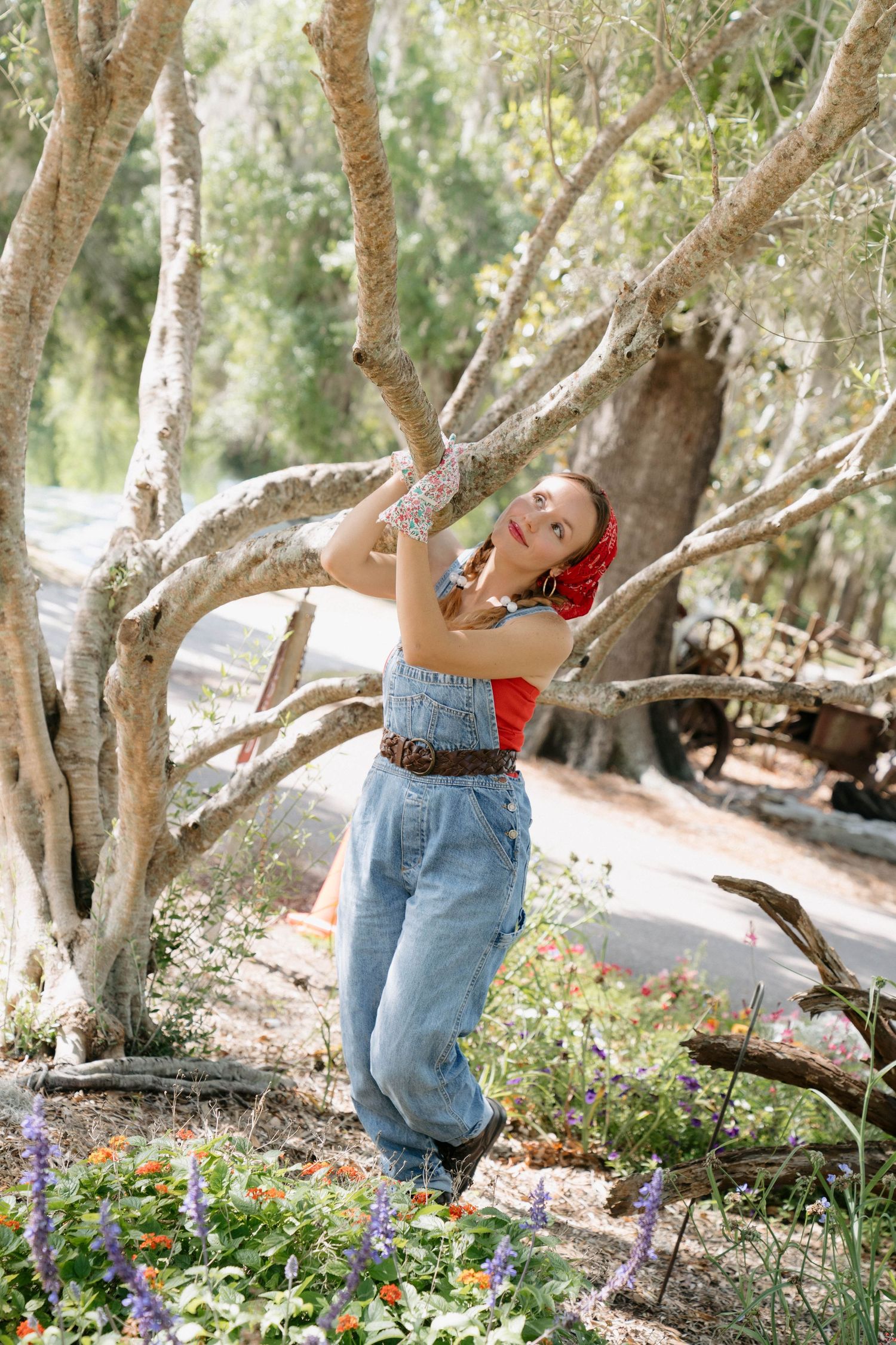 Young woman in denim overalls and red top holds tree branch surrounded by colorful wildflowers in a sunlit garden.
