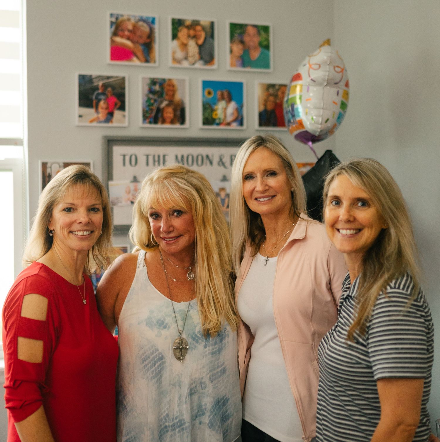 Four women pose together in a home with framed photos and a balloon decoration visible in the background.