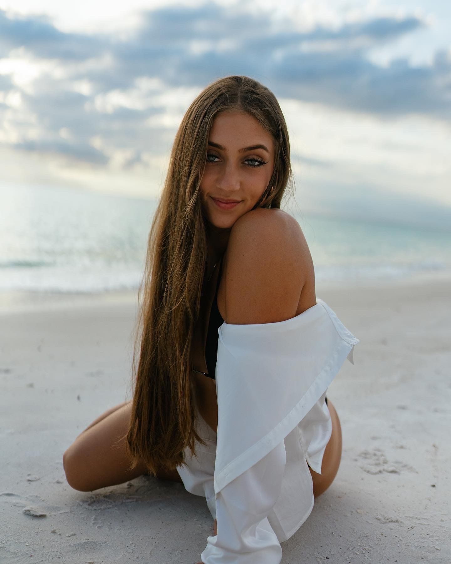Figure in a white flowy top poses on a sandy beach at sunset with clouds and ocean in the background.