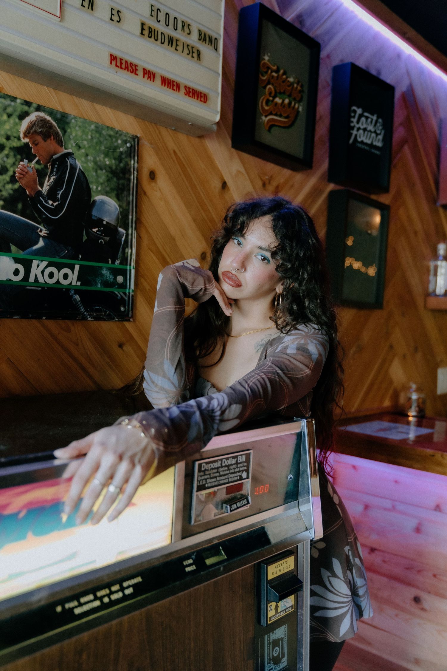 A person in a gray sweater leans over a vintage pinball machine in a dimly lit arcade with neon purple lighting.