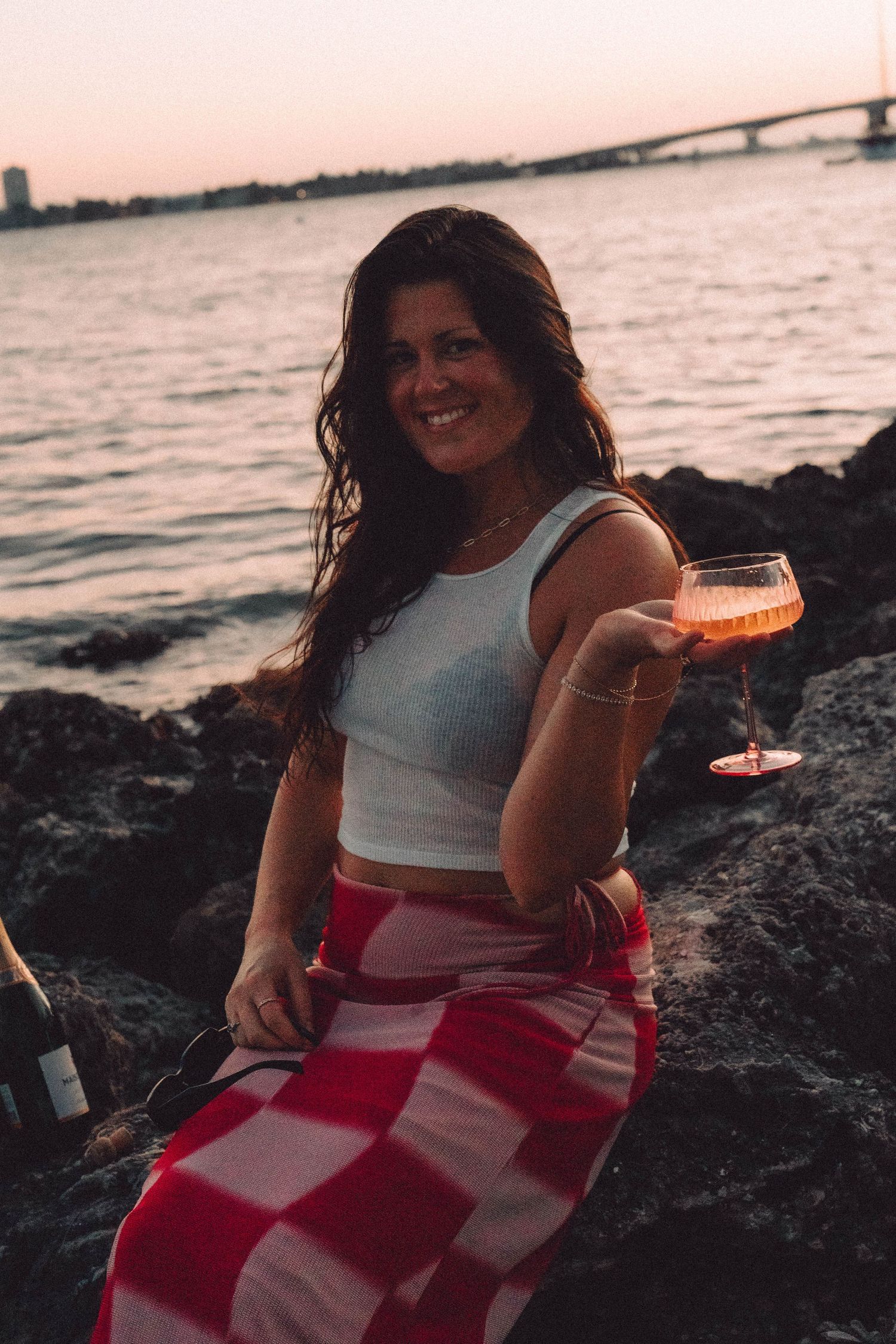 A smiling brunette woman holding a pink cocktail while sitting on rocks by the water at sunset.