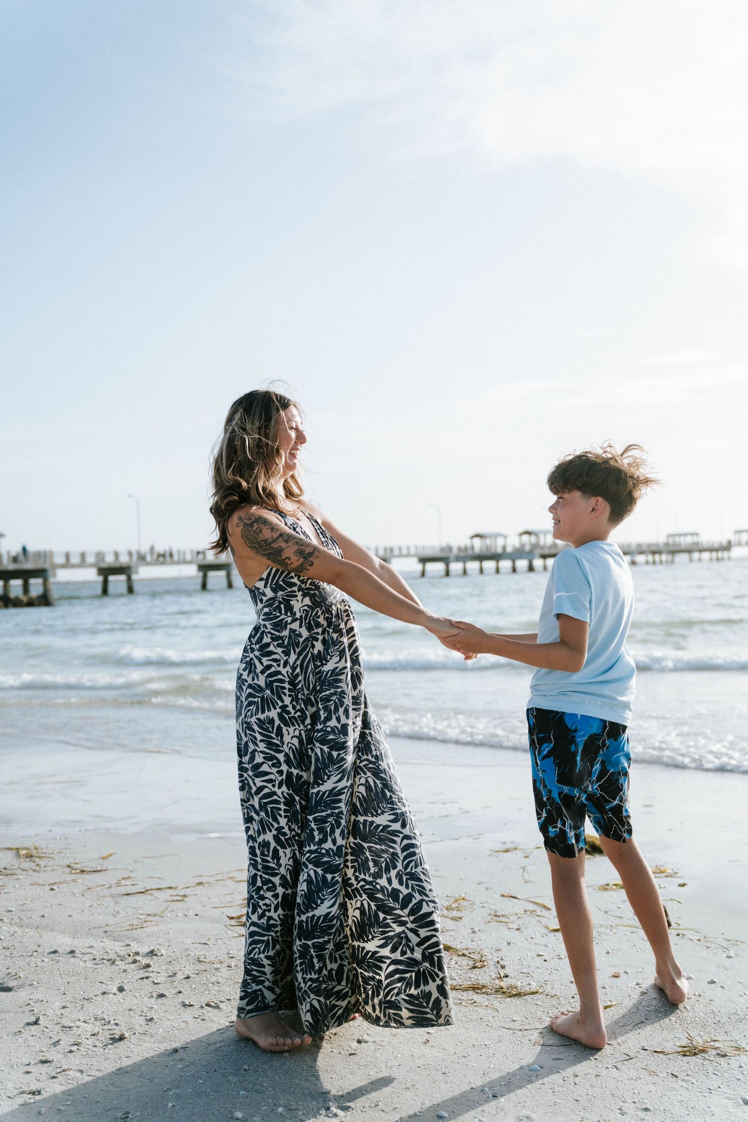 A mother and son share a joyful moment dancing together on a beach with a pier in the background.
