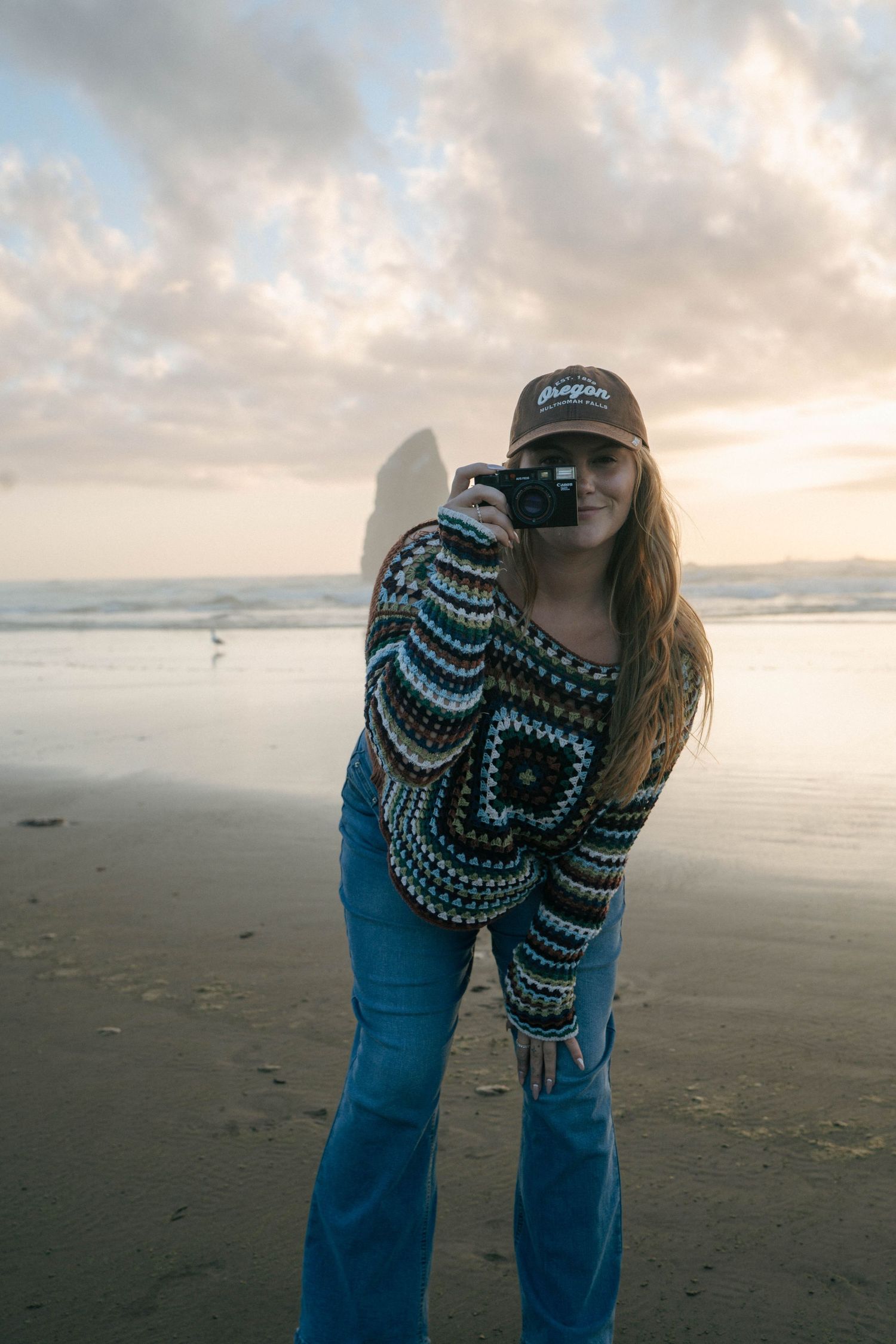 A person in a patterned sweater and jeans bends down to explore the sandy beach at sunset with cloudy skies.