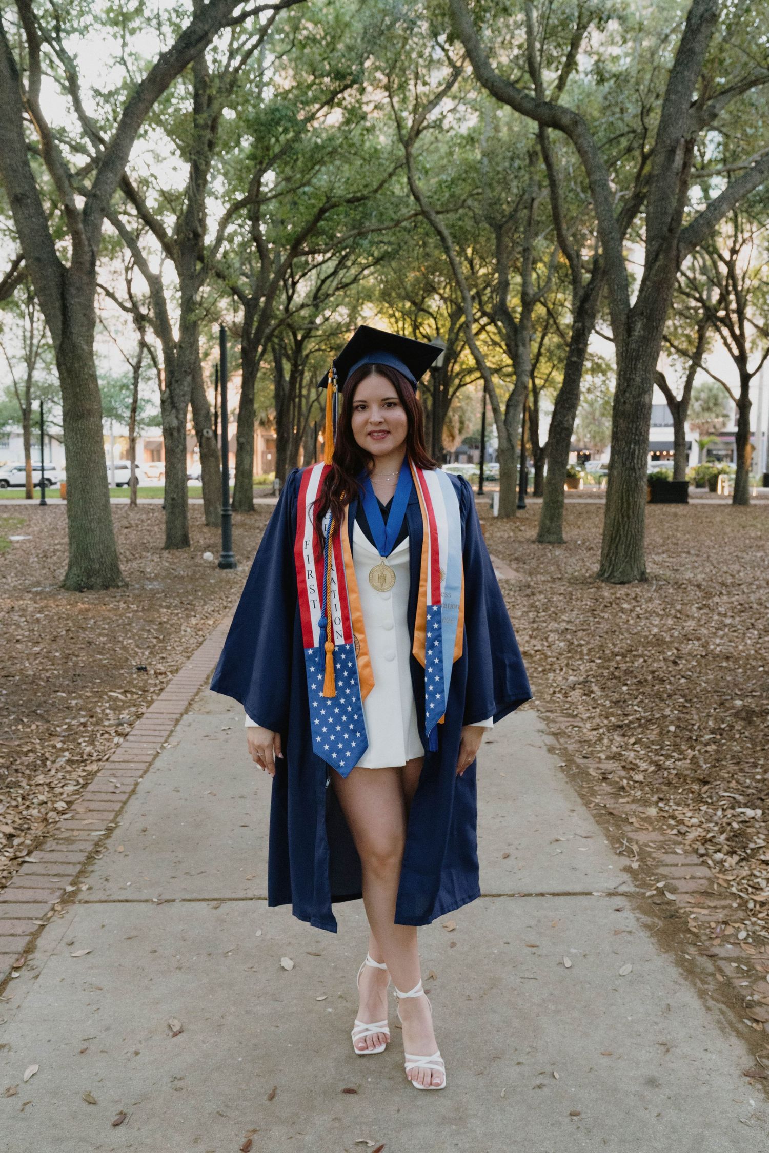 Young woman in navy graduation cap and gown with honor stoles walks along a tree-lined campus pathway.