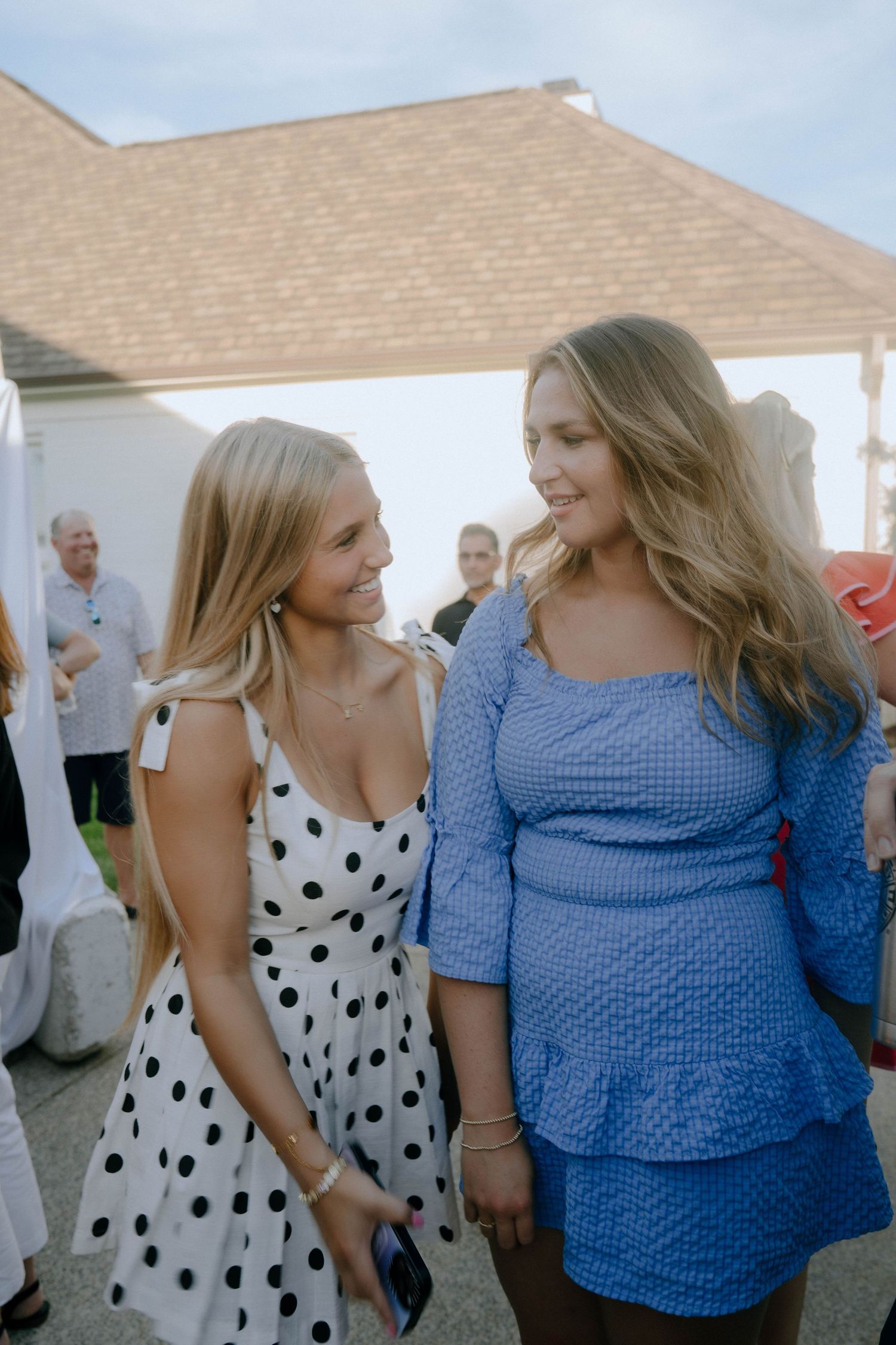 Two women in casual dresses share a conversation at an outdoor gathering, one wearing polka dots and one in blue lace.