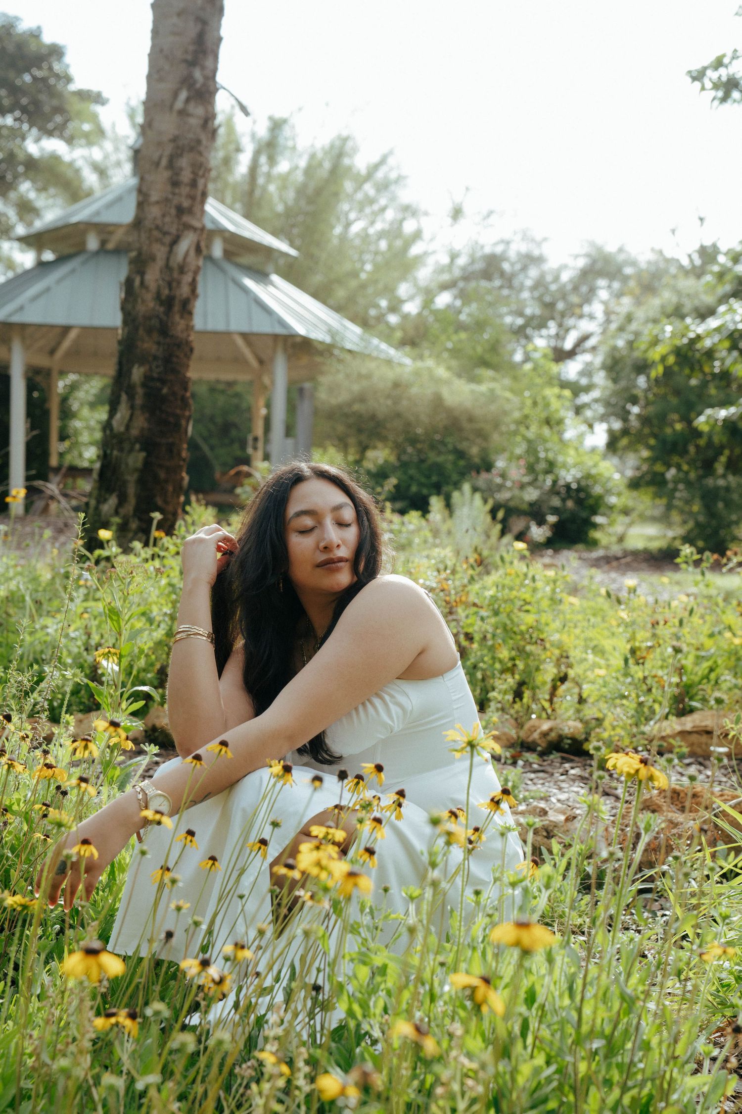 A figure in a white dress sits gracefully among yellow wildflowers in a garden with a gazebo in the background.