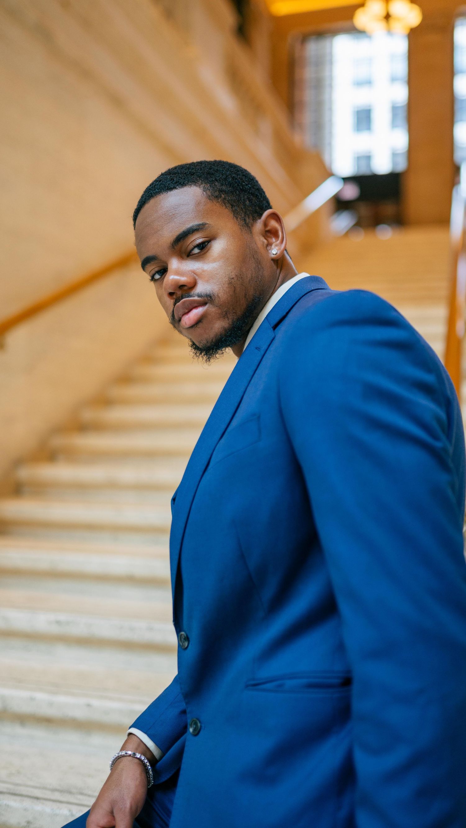 Professional portrait of someone in a sharp royal blue suit jacket posed on marble stairs in warm lighting.