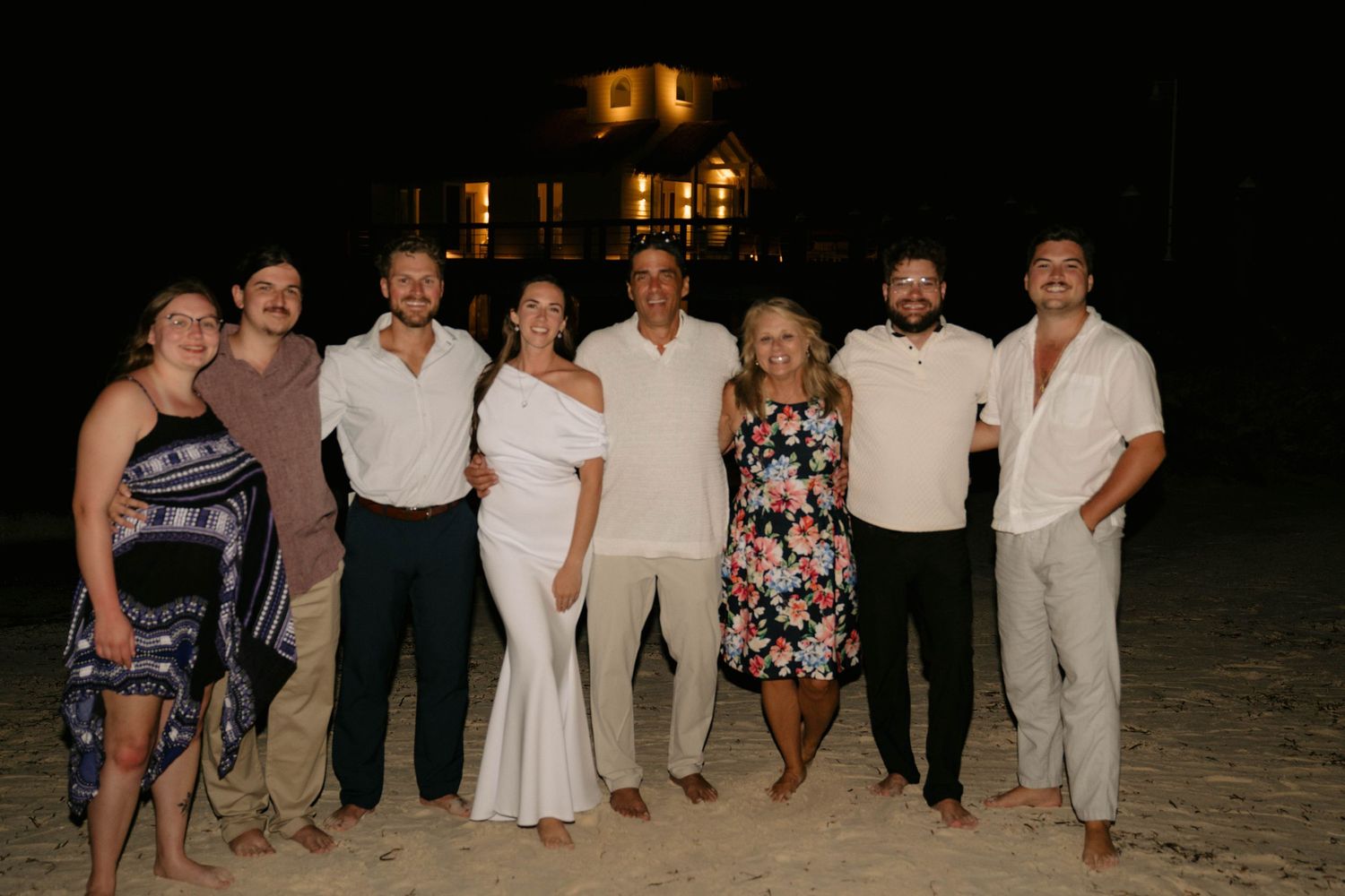 Group of eight adults posing together on a beach at night with illuminated building in background.