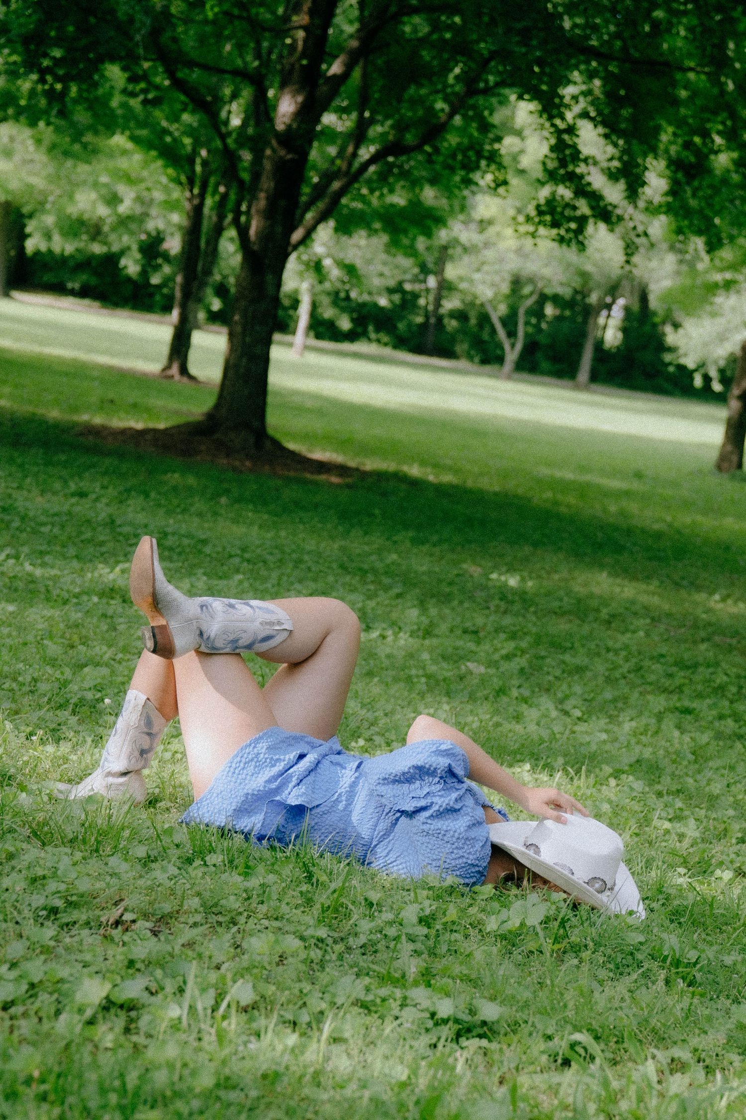 Woman in blue dress and cowboy boots lying on green grass in a park, hat covering her face on a sunny day.