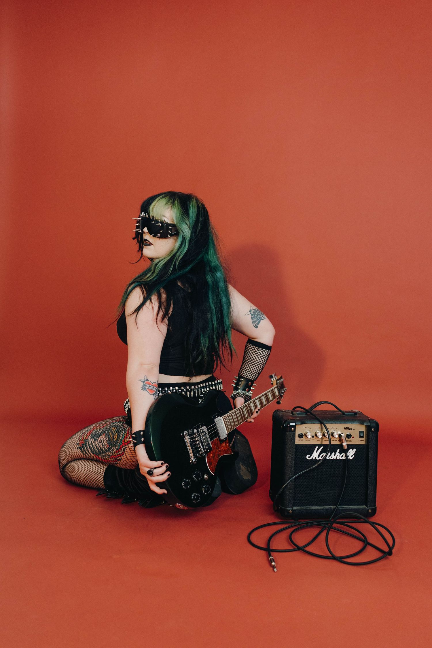 Female rock guitarist with green hair and tattoos poses with black electric guitar beside Marshall amp on red background.