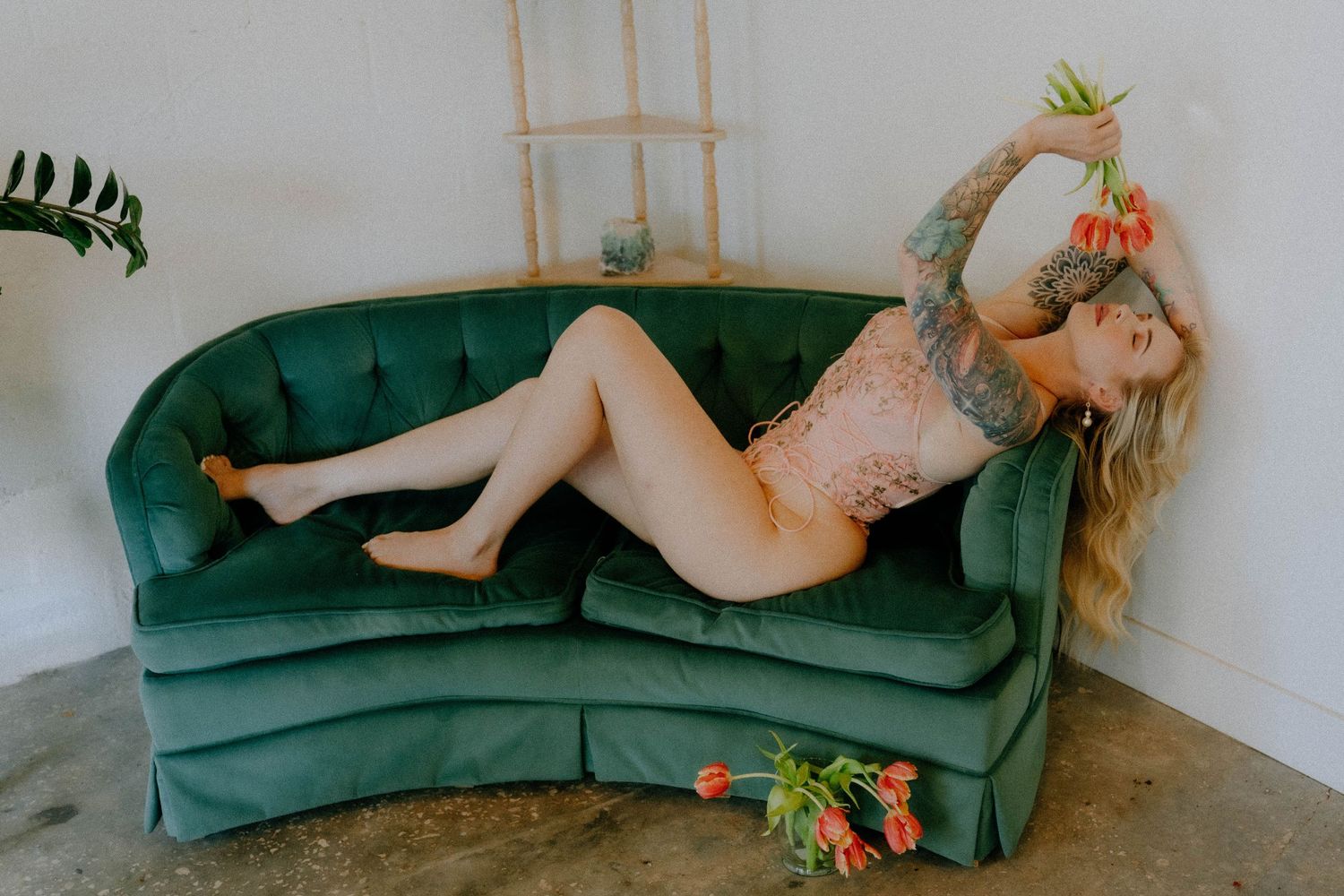 A person in lingerie reclines on an emerald green velvet couch with scattered orange lilies.