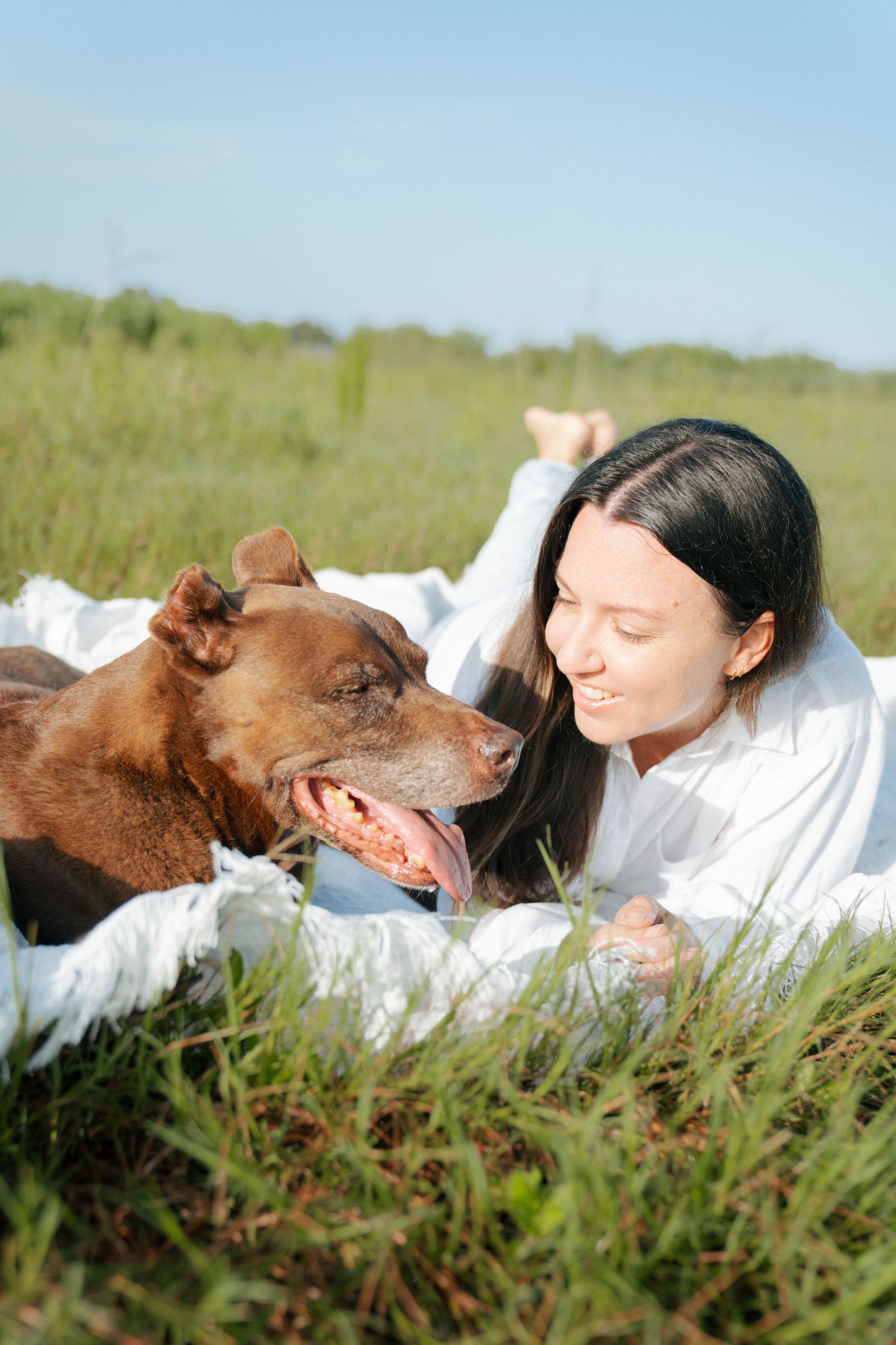 Young woman lying on white blanket in green field, smiling face-to-face with happy brown dog on sunny day.