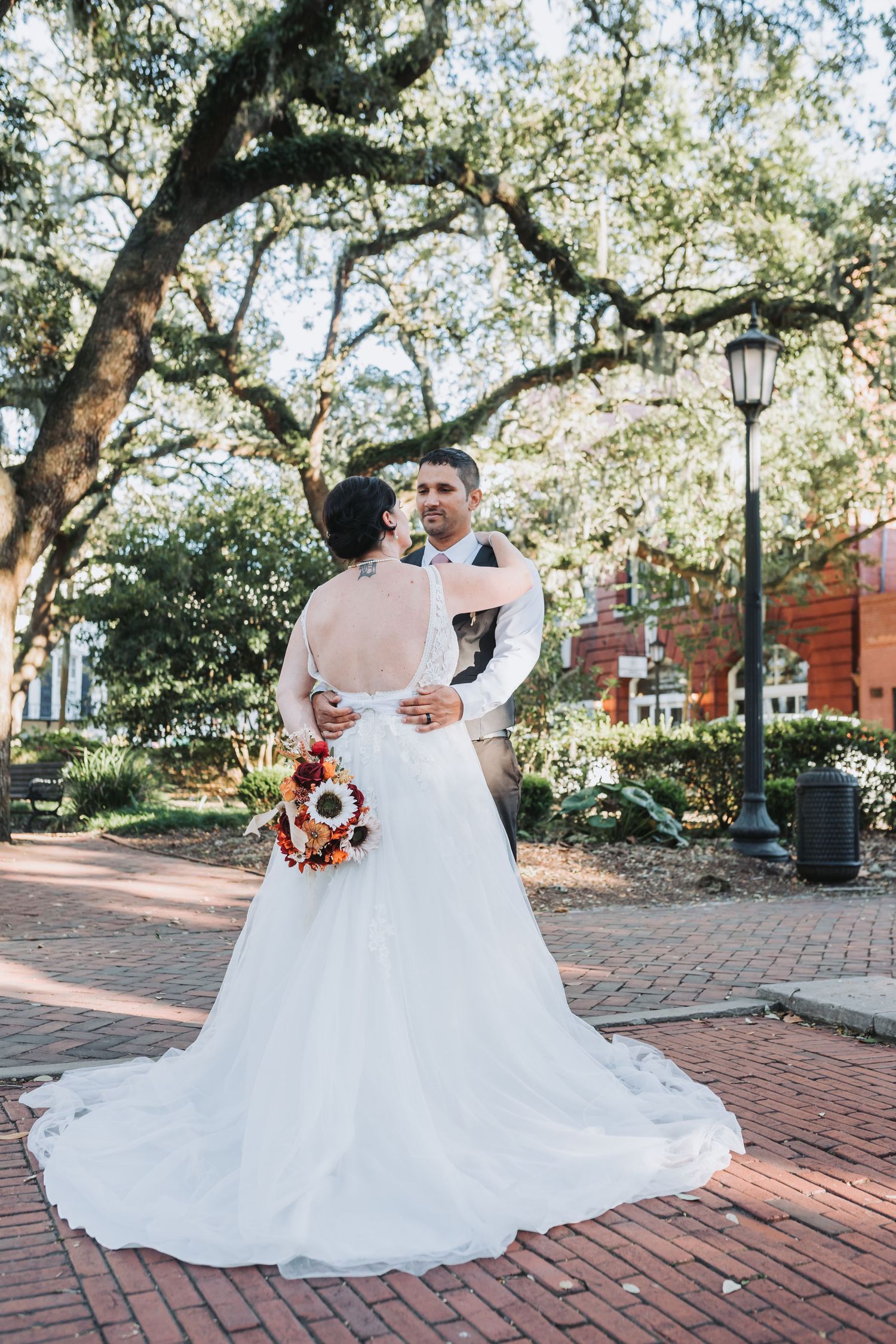 Wedding couple embracing on brick pathway under oak trees with trailing white dress and autumn bouquet.