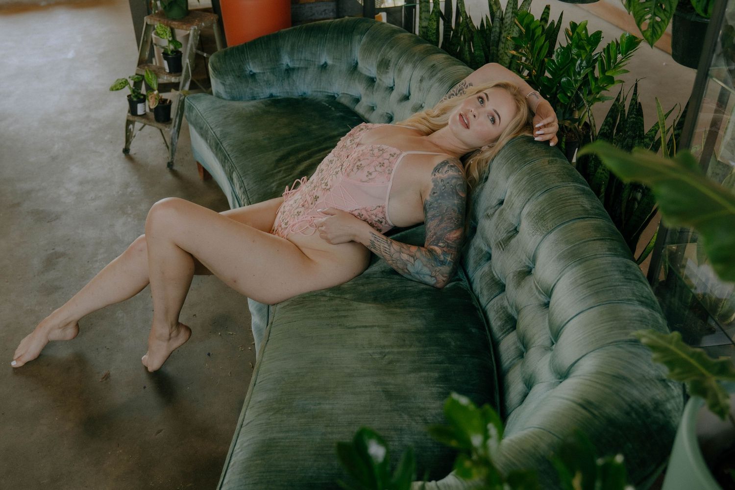 Someone poses on a green velvet tufted couch surrounded by potted plants in a boudoir photography session.